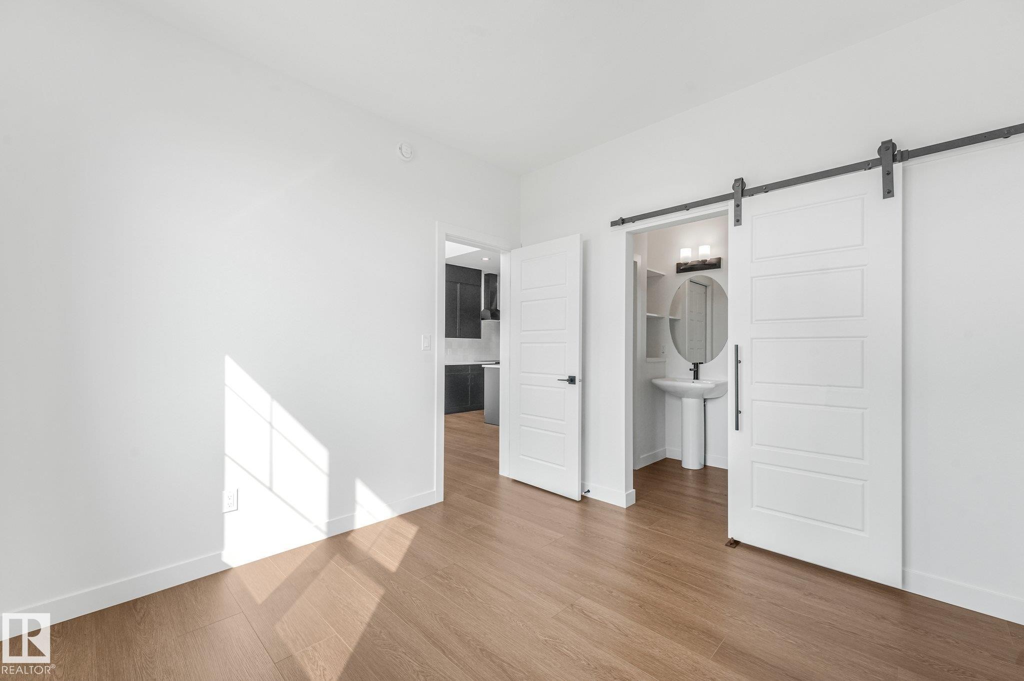 Wood-finish flooring, white walls, and a barn door leading to a bathroom featuring a pedestal sink, round mirror, and vanity light - 511 157 Avenue Ne, Edmonton, AB