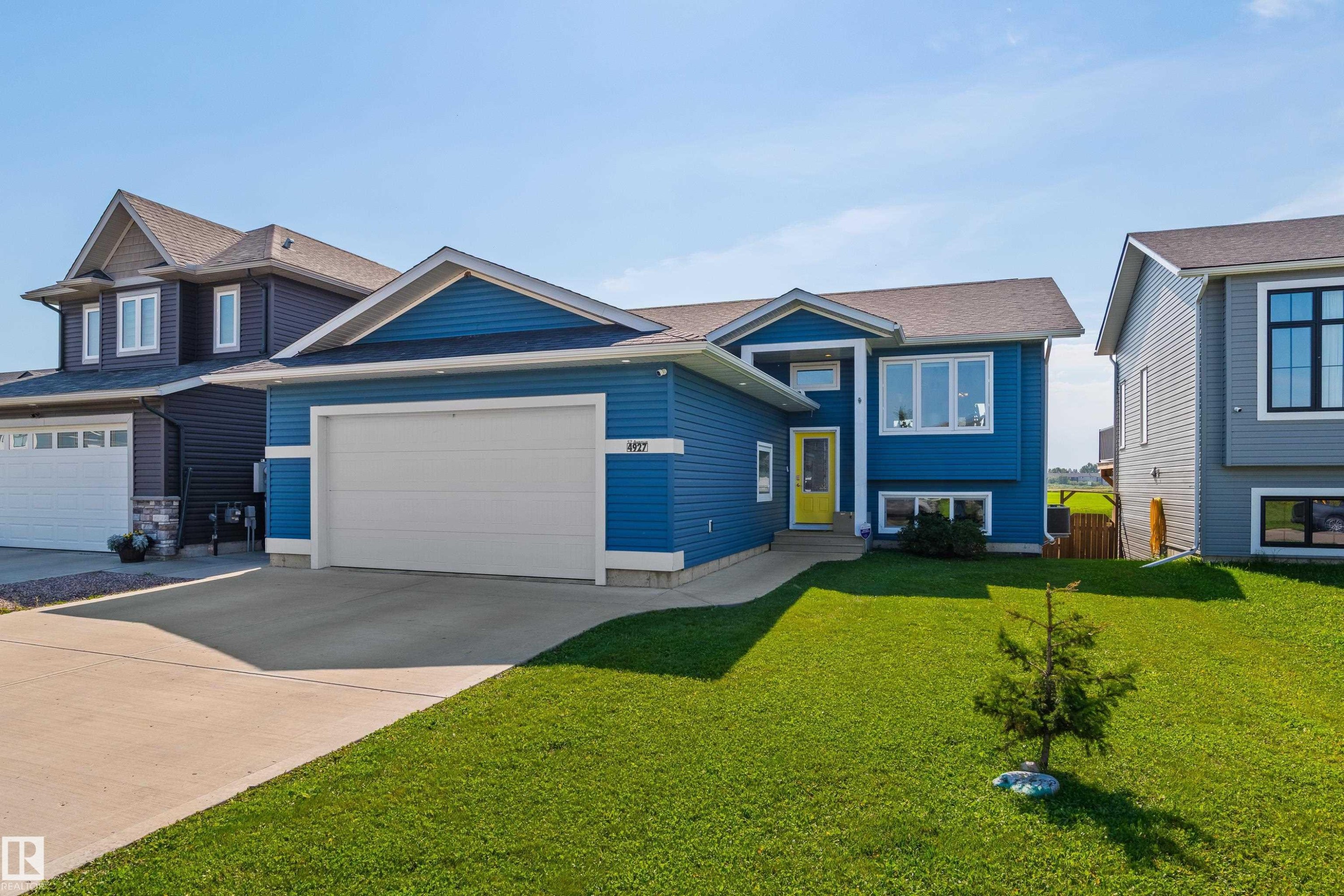 Blue horizontal siding exterior with white trim, attached garage, concrete driveway, and a front lawn with a single sapling - 4927 57 Avenue, Cold Lake, AB
