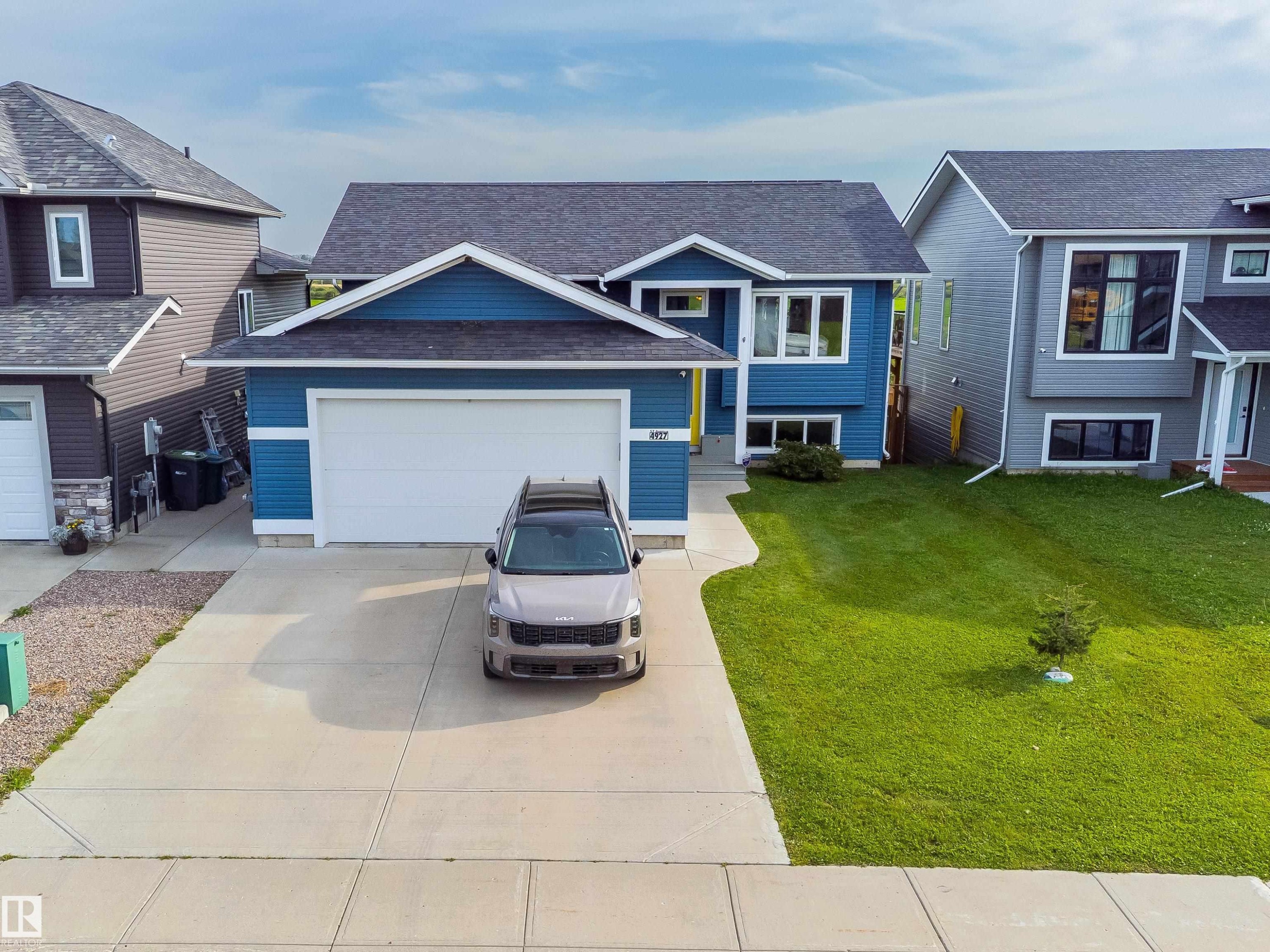 Contemporary residential structure featuring a blue exterior, attached garage, concrete driveway, and a front lawn with a young tree - 4927 57 Avenue, Cold Lake, AB