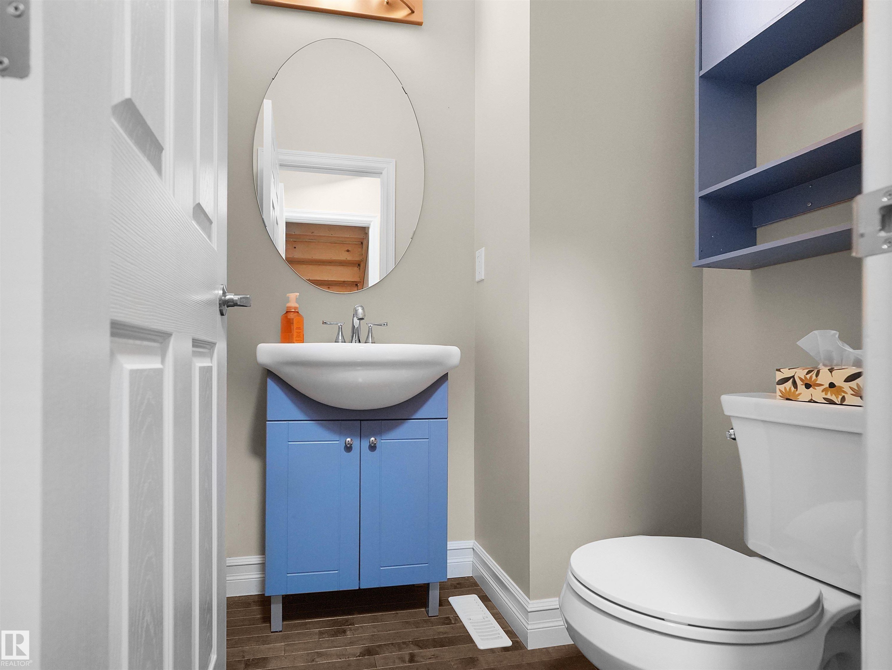 Pedestal sink with blue vanity, oval wall mirror, wall-mounted shelving, and wood-finish flooring - 5190 1B Avenue Sw, Edmonton, AB