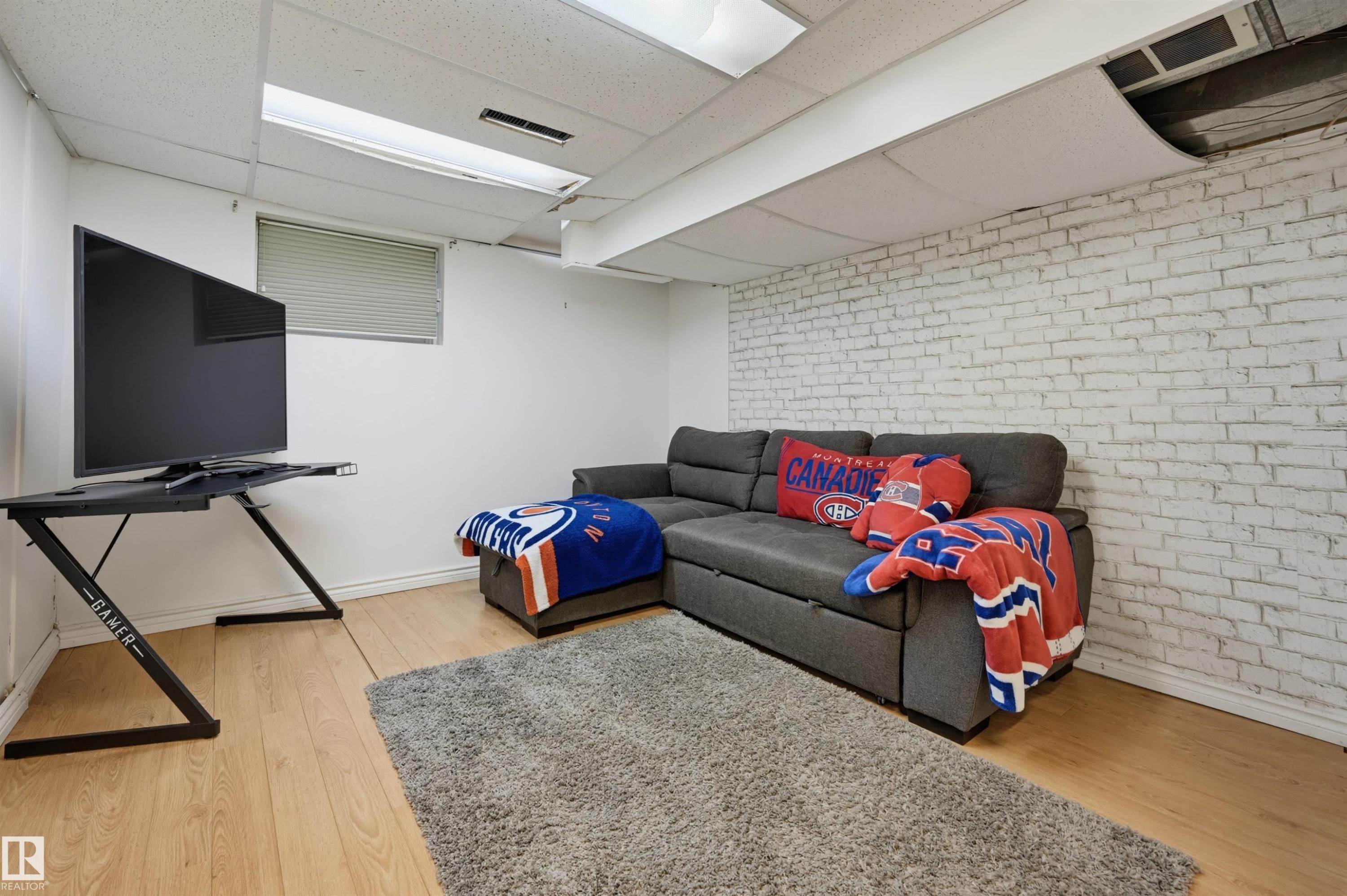 White painted brick accent wall, wood-finish flooring, drop ceiling with integrated lighting, and window with blinds - 10726 128 Street Nw, Edmonton, AB