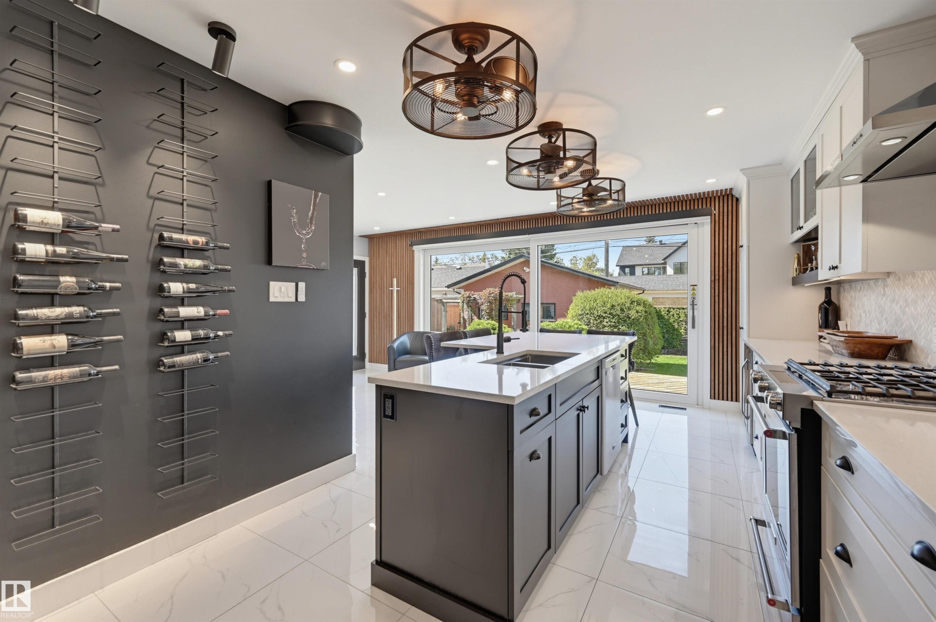 Gleaming tile flooring, a central kitchen island with integrated sink, white cabinetry with dark hardware, a stainless steel gas range, and a glass sliding door leading to the exterior - 10726 128 Street Nw, Edmonton, AB