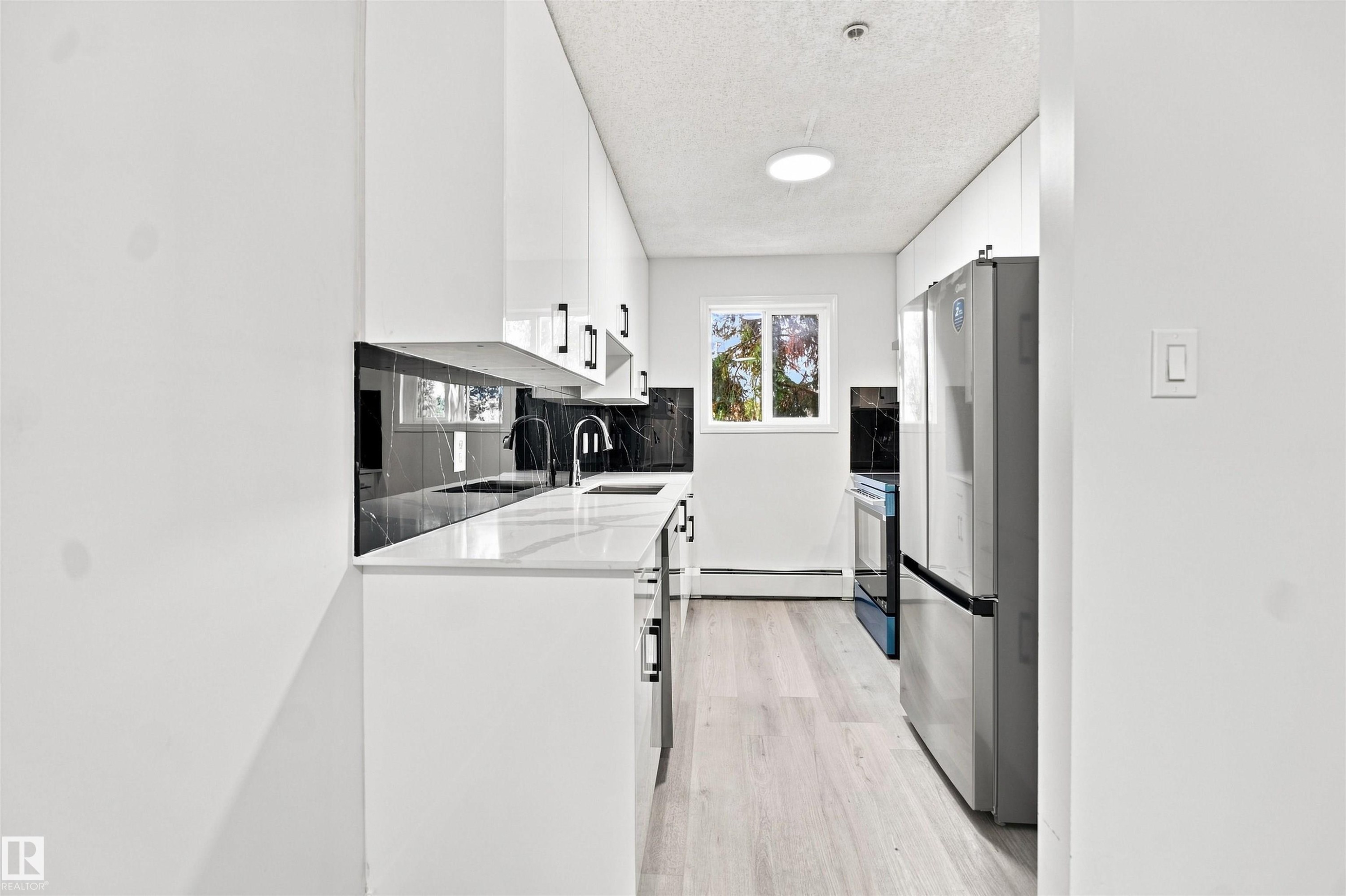 White cabinetry with black hardware, black marble-patterned backsplash, white countertops, stainless steel appliances, and light wood-finish flooring - #124 10511 42 Av Nw, Edmonton, AB