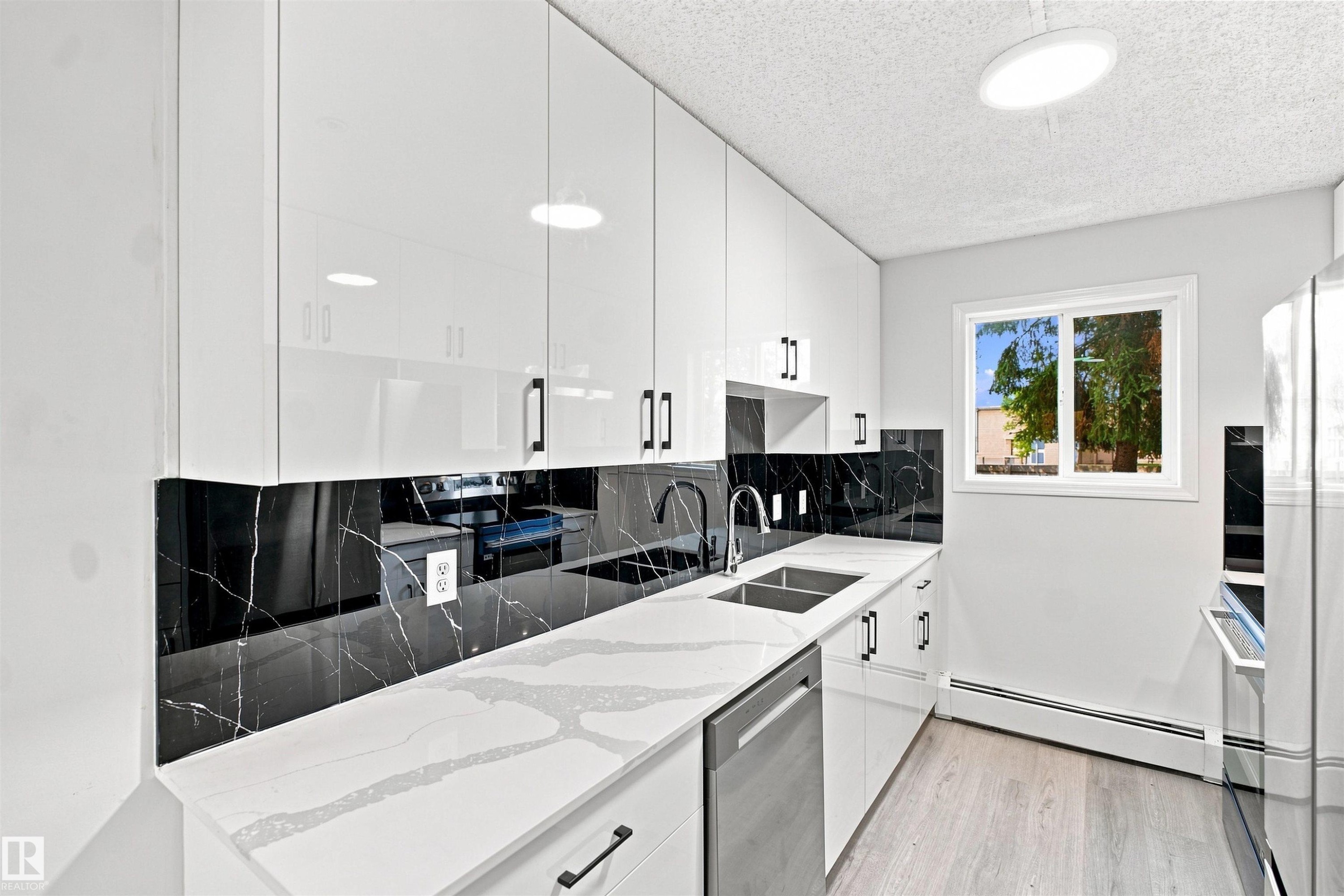White high-gloss cabinetry, black and white veined backsplash, white veined countertop, stainless steel double basin sink, and light wood-finish flooring - #124 10511 42 Av Nw, Edmonton, AB