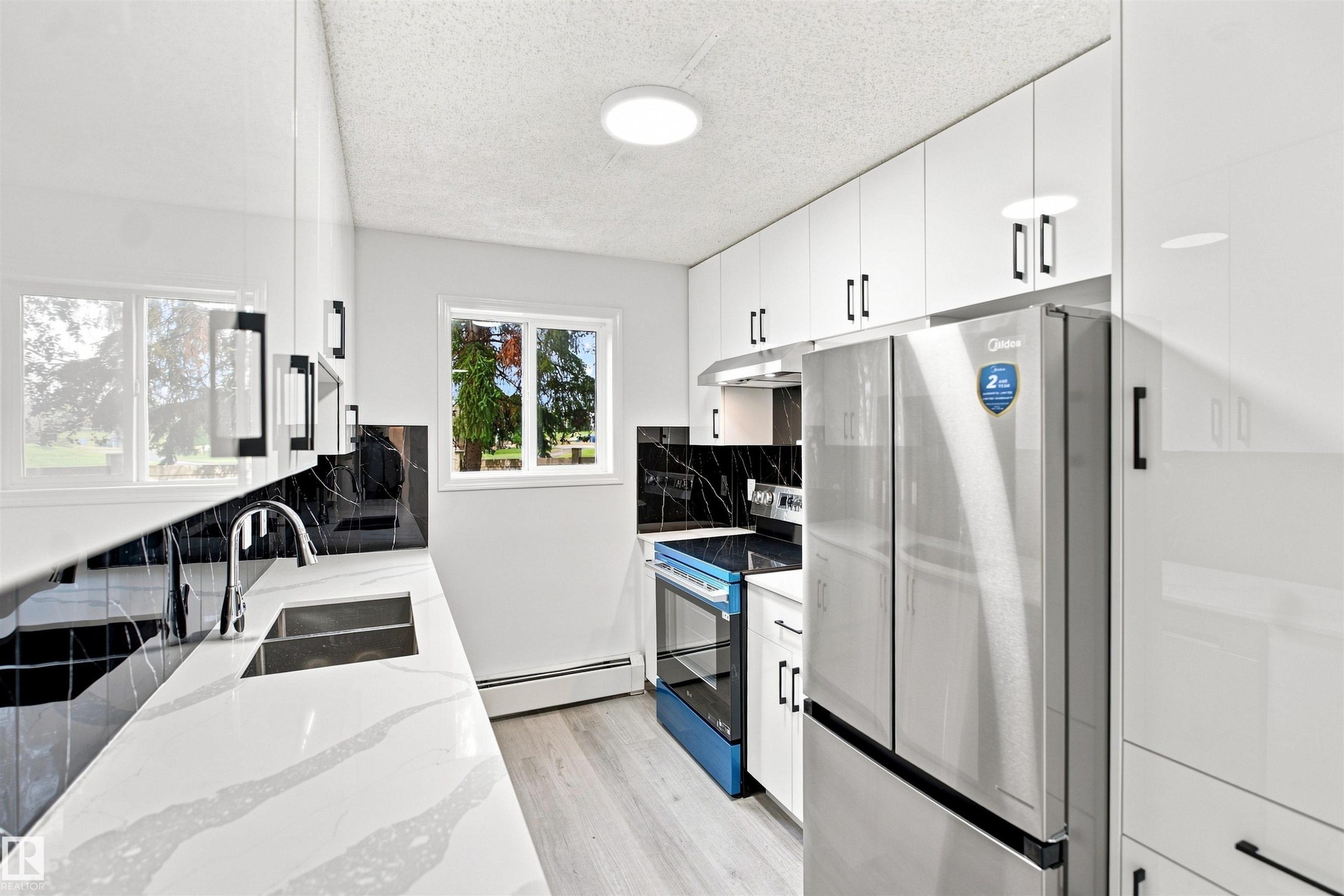 White cabinetry with black hardware, white countertops, black-patterned backsplash, stainless steel appliances, and light wood-finish flooring - #124 10511 42 Av Nw, Edmonton, AB