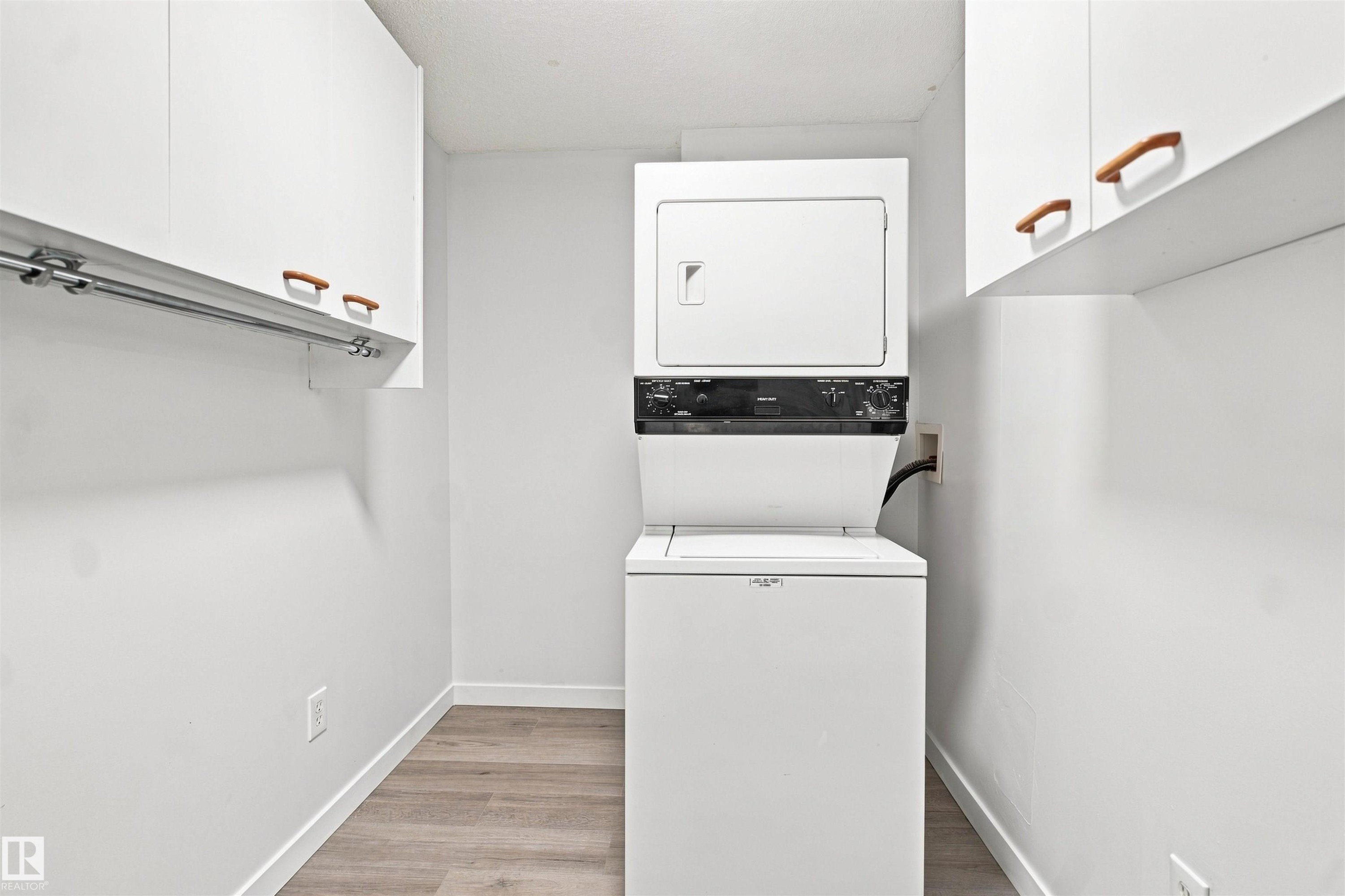 Laundry area with stackable appliances, wood-finish flooring, and white cabinetry featuring wood pulls - #124 10511 42 Av Nw, Edmonton, AB