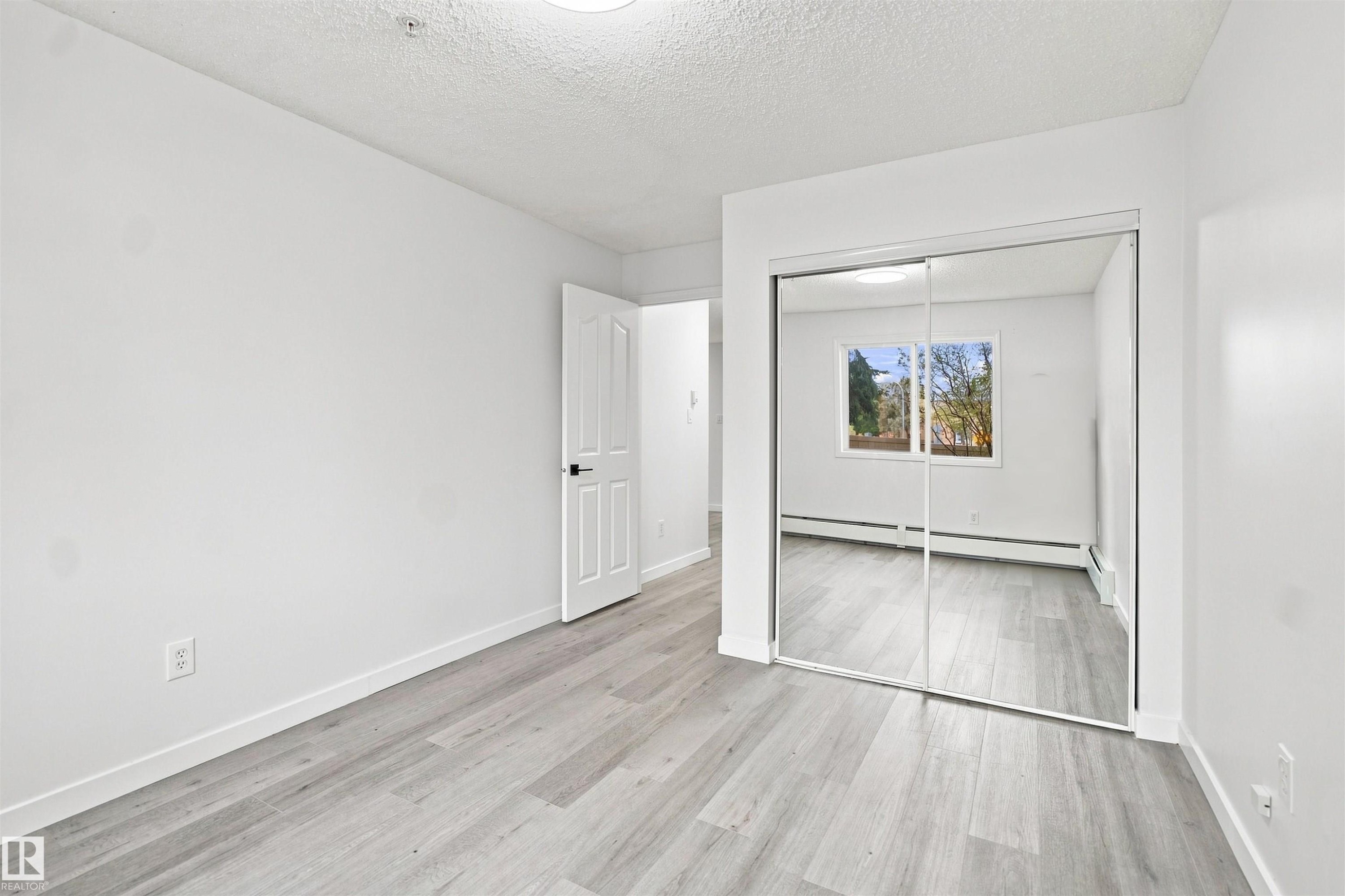 Room featuring wood-finish flooring, a white paneled door with dark hardware, and a mirrored sliding closet door - #124 10511 42 Av Nw, Edmonton, AB