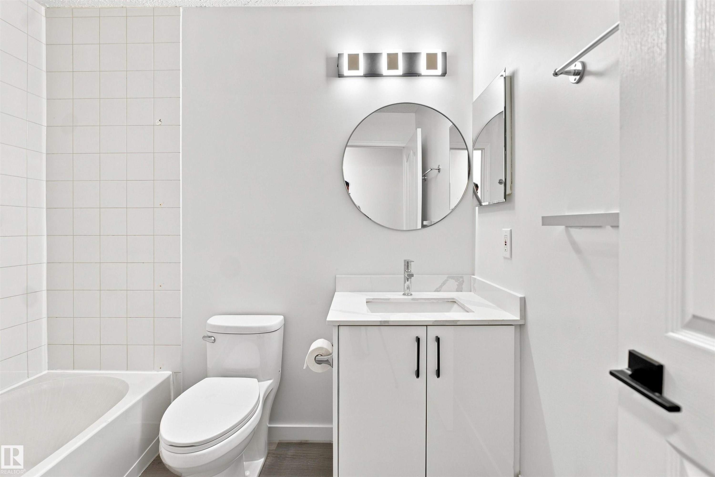 White tiled shower with a bathtub, white vanity with an integrated sink, and a circular mirror - #124 10511 42 Av Nw, Edmonton, AB