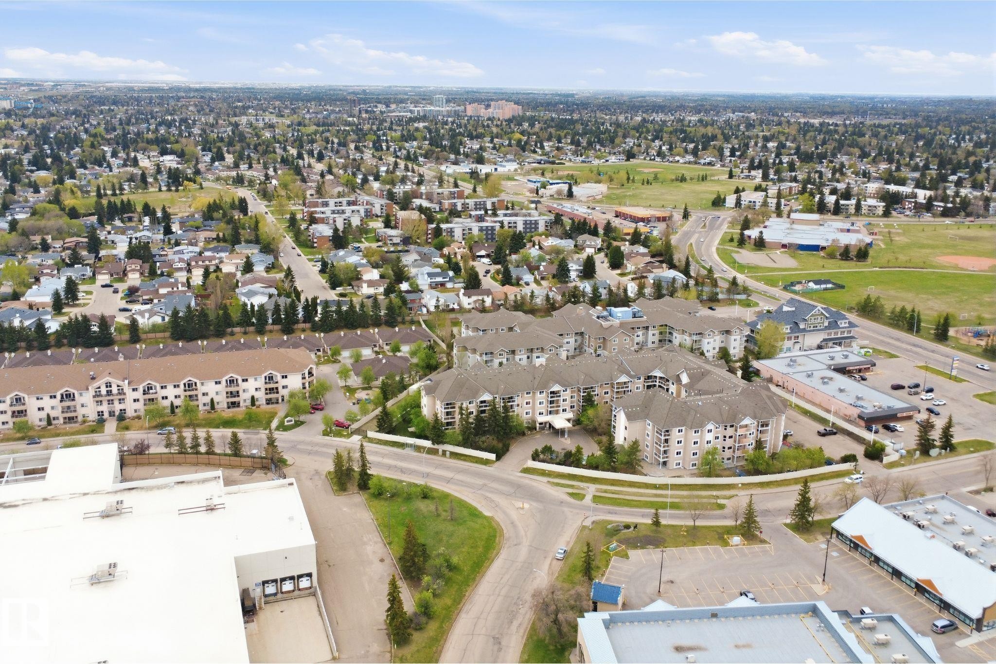 Expansive aerial perspective showcasing a developed neighborhood with mature trees, multi-story residential buildings, commercial structures, and paved roadways - #124 10511 42 Av Nw, Edmonton, AB