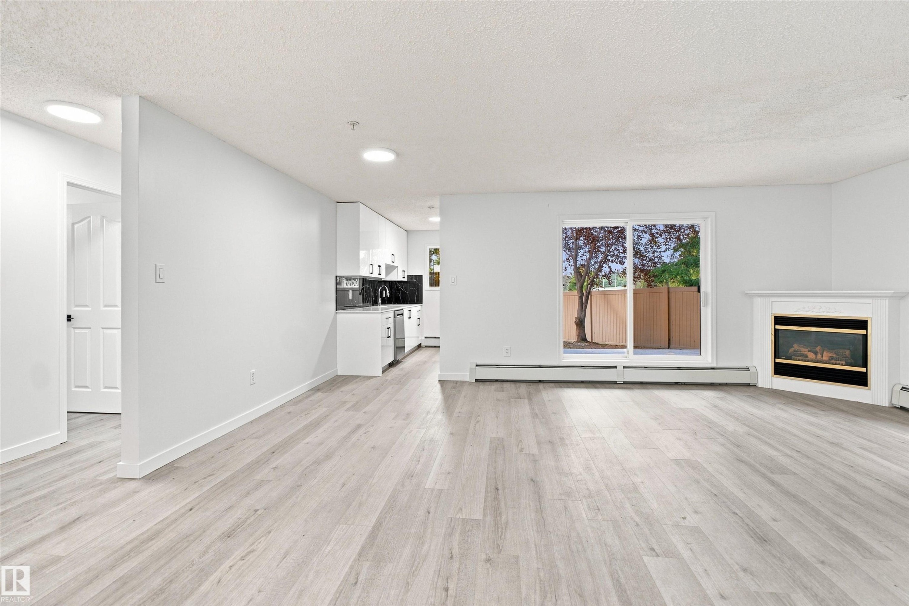Open-concept living space with wood-finish flooring, a white mantel fireplace, and a sliding glass door - #124 10511 42 Av Nw, Edmonton, AB