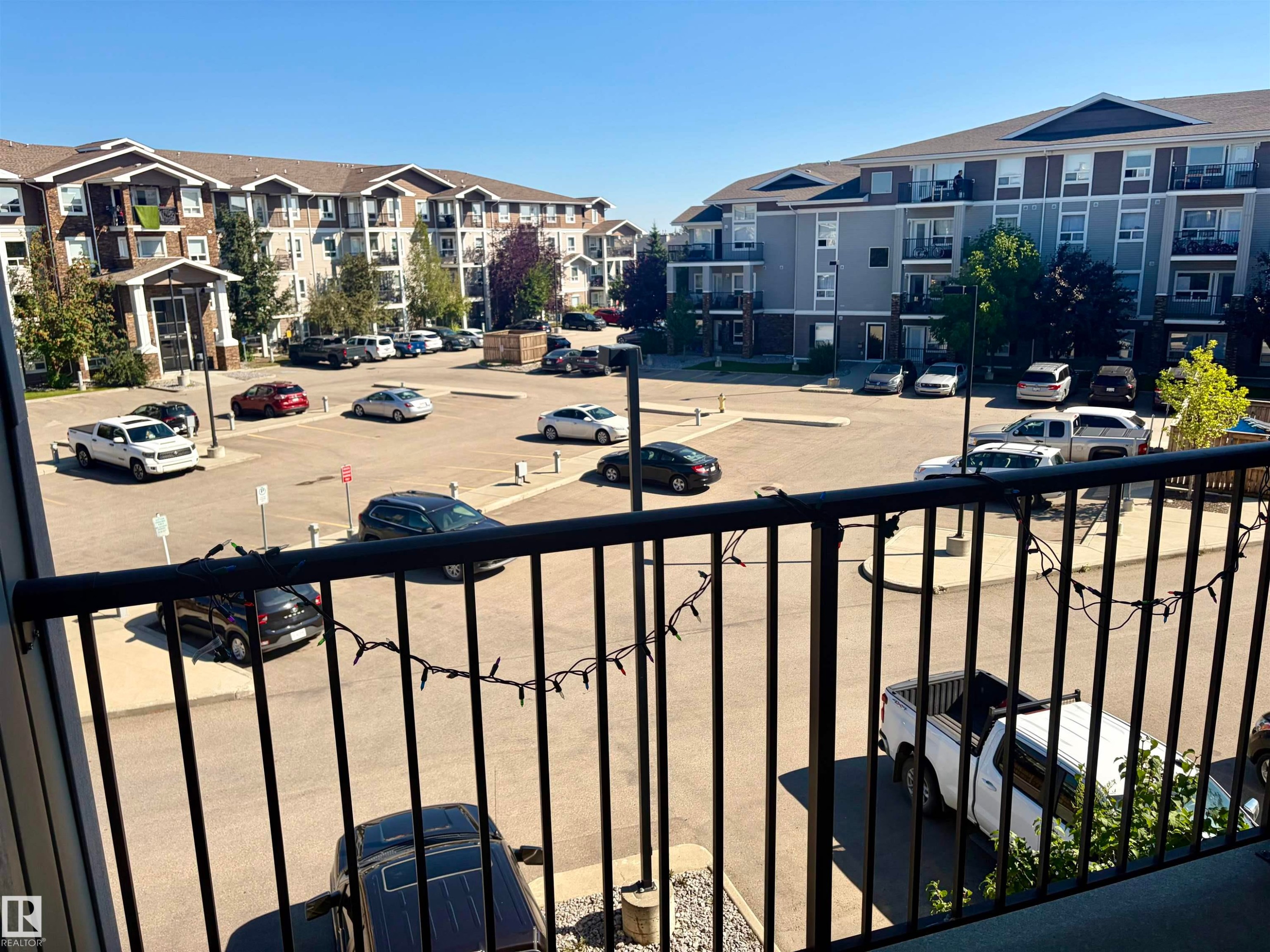 Open-air balcony with black metal railings overlooking a paved parking area and multi-story residential buildings featuring exterior balconies - #1304 9363 Simpson Dr Nw, Edmonton, AB
