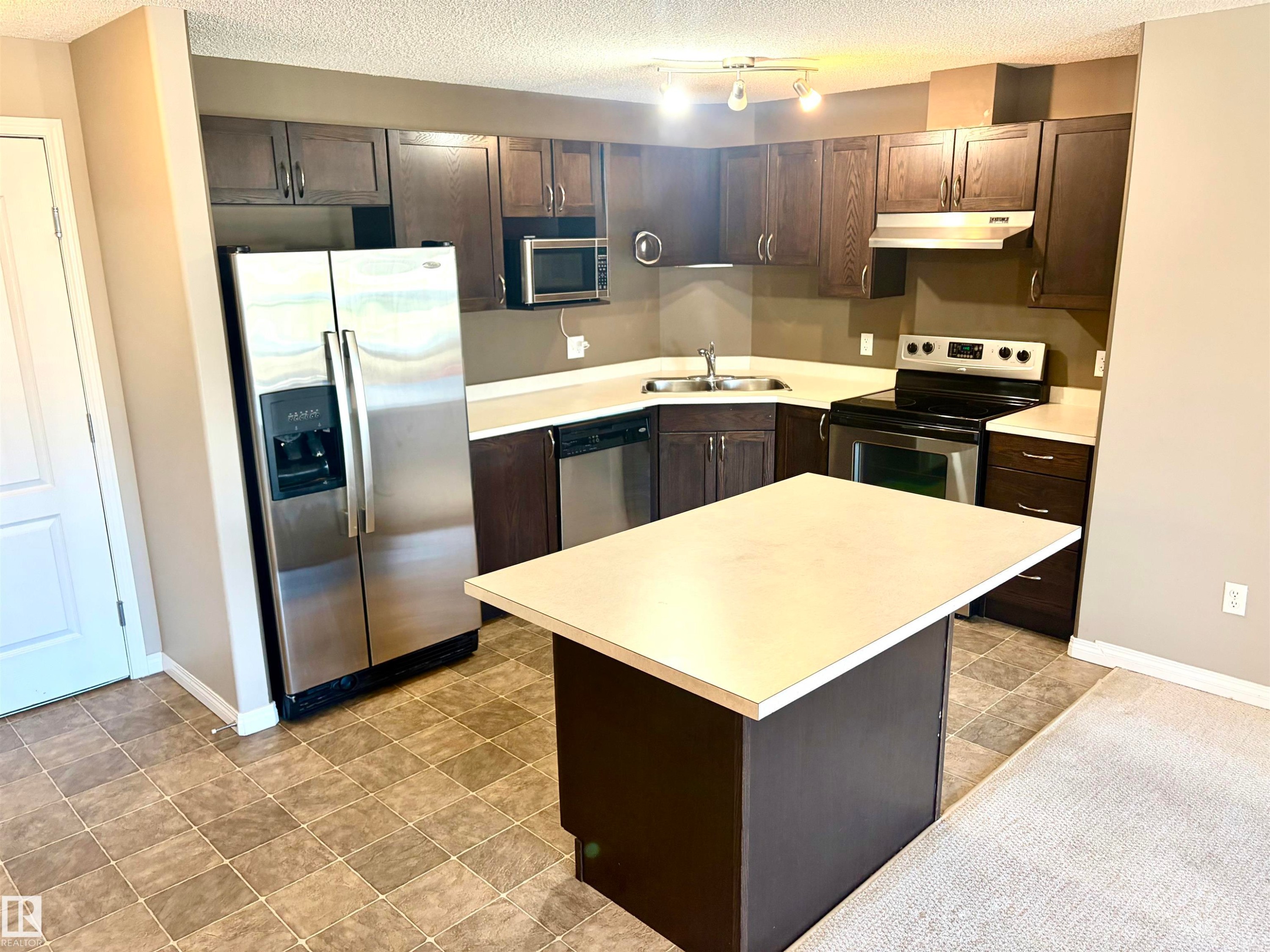 Kitchen featuring stainless steel appliances, dark wood-finish cabinetry, a built-in island, track lighting, and tiled flooring - #1304 9363 Simpson Dr Nw, Edmonton, AB