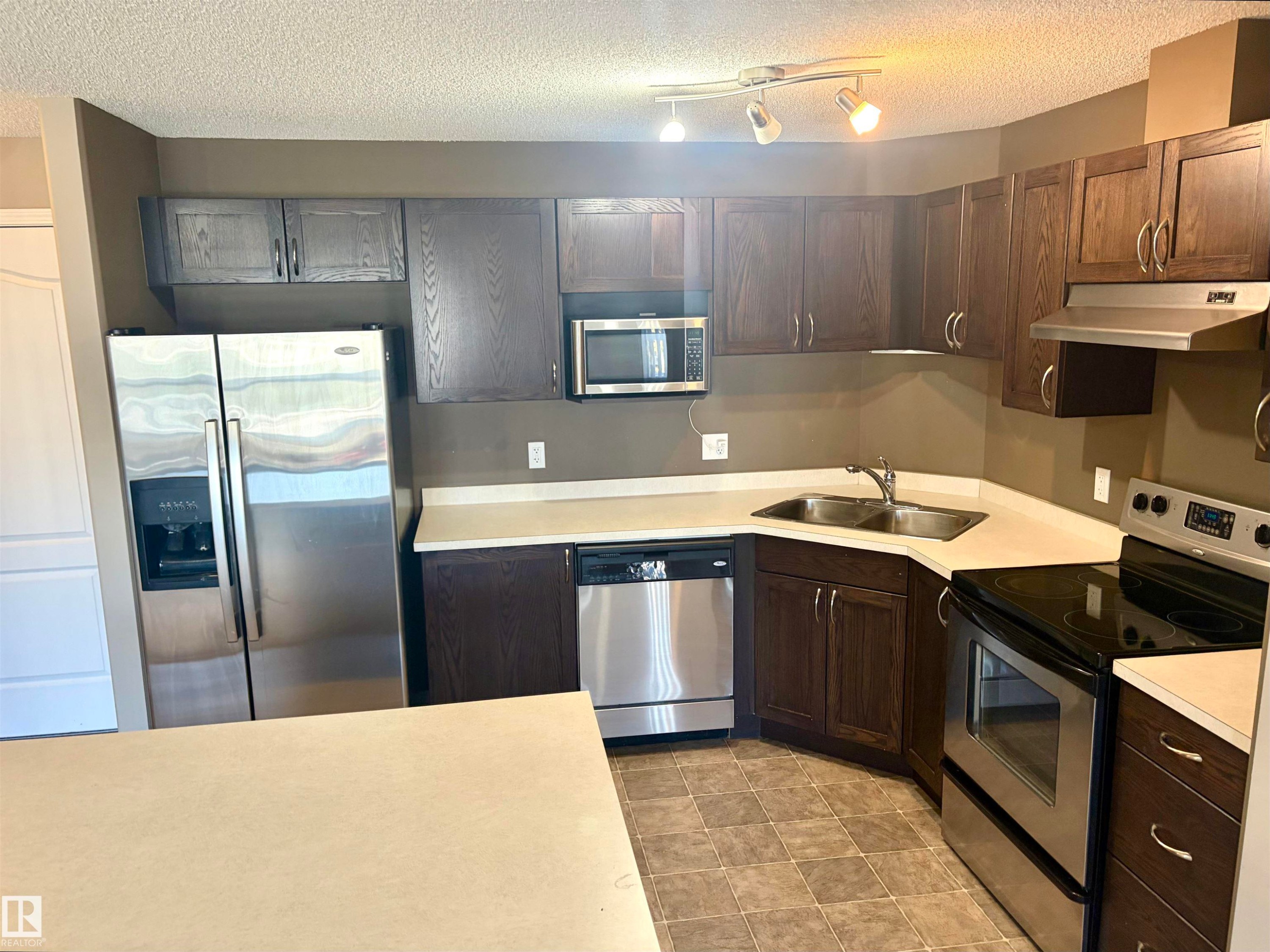 Kitchen featuring wood cabinetry, stainless steel appliances, light-toned countertops, and track lighting - #1304 9363 Simpson Dr Nw, Edmonton, AB