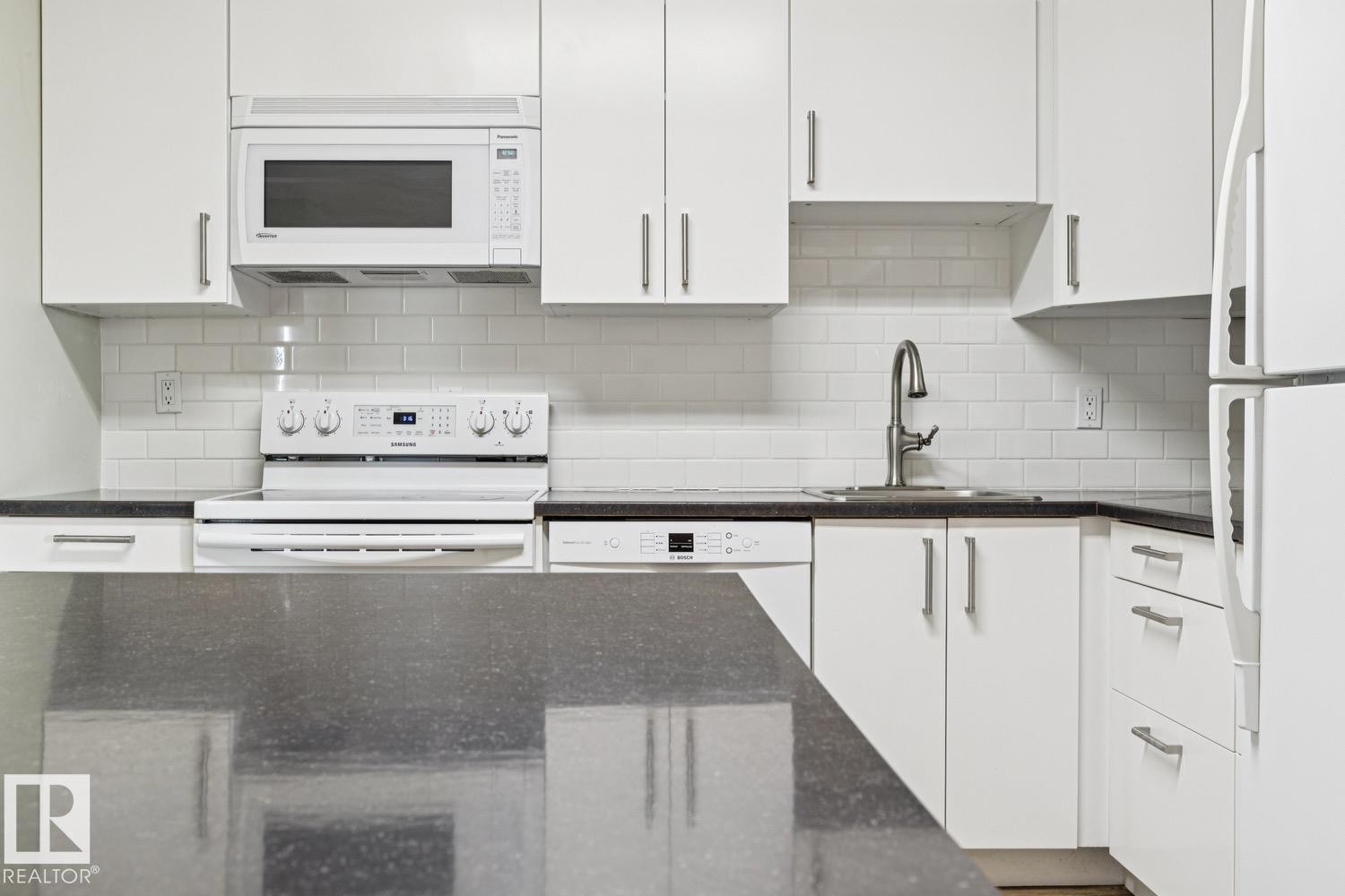 White cabinetry, dark stone countertops, white subway tile backsplash, stainless steel sink with gooseneck faucet, and white appliances - #905 9929 113 St Nw, Edmonton, AB