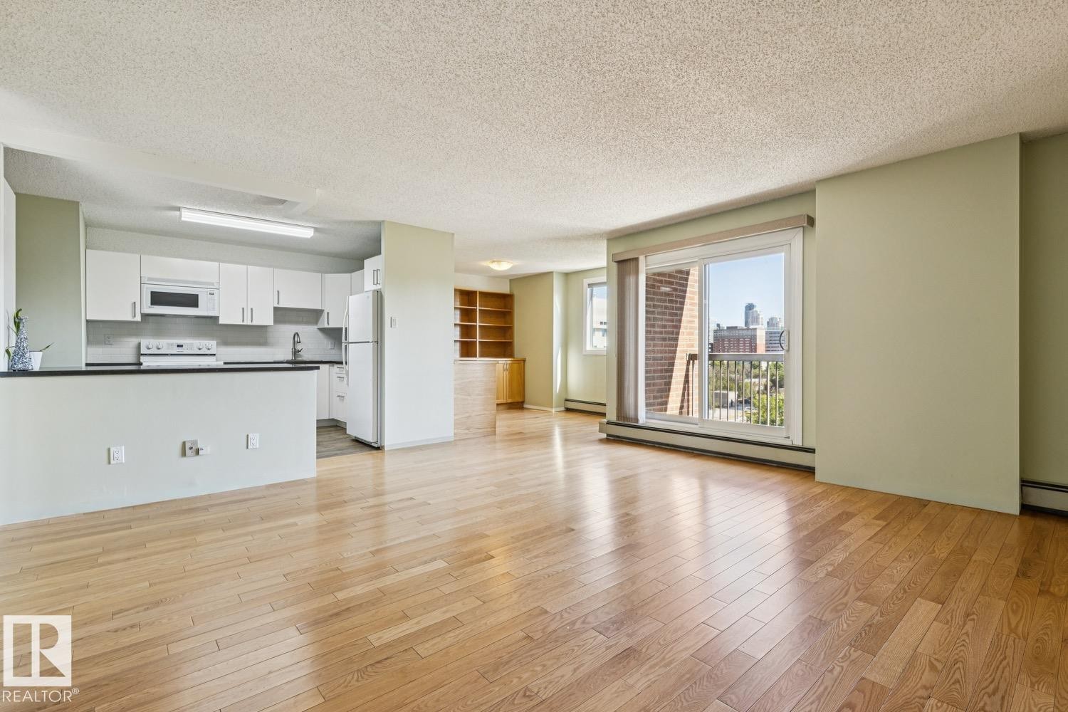 Open-concept living space featuring wood-finish flooring, a kitchen with white cabinetry and dark countertops, built-in shelving, and a sliding glass door leading to a balcony - #905 9929 113 St Nw, Edmonton, AB