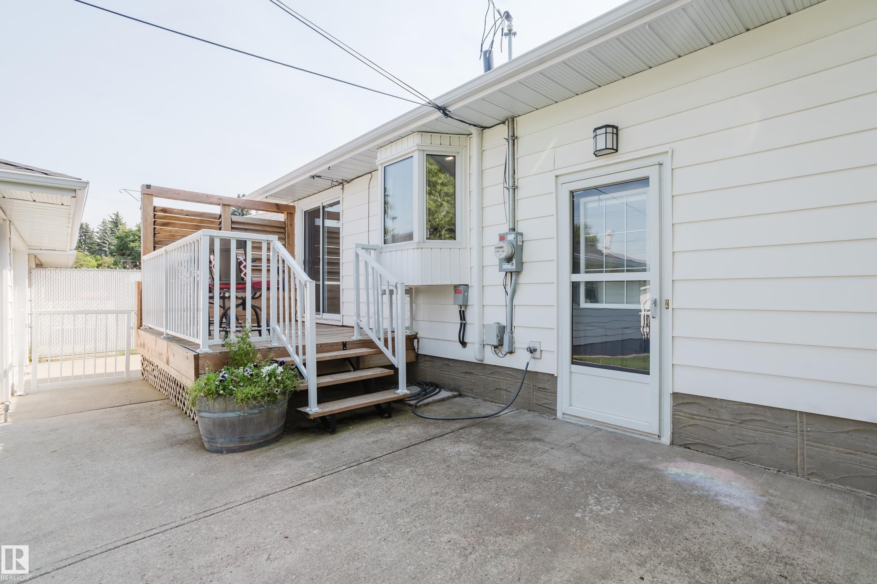 Exterior white siding with a ground-level concrete patio, a raised wood deck with white railings, and a glass-paneled exterior door - 4708 106 Avenue Nw, Edmonton, AB