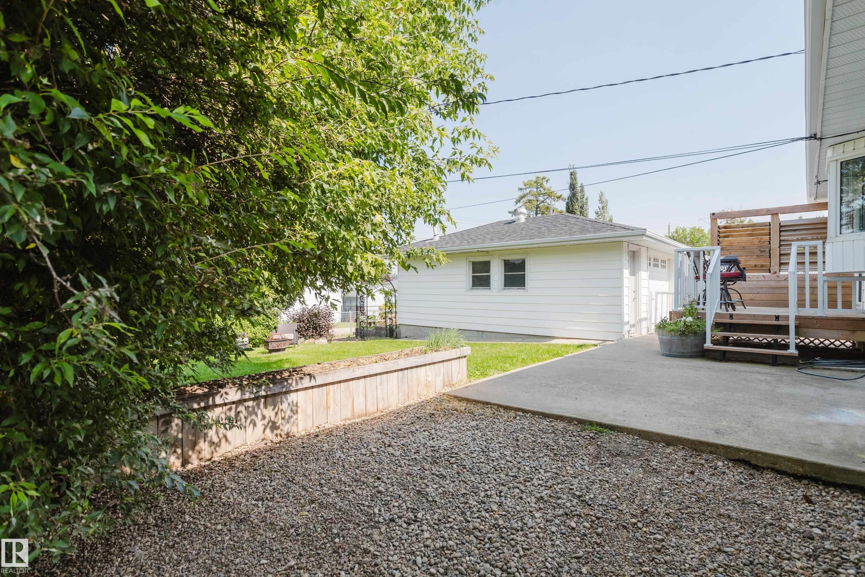 Gravel parking area with a wood-retaining wall, leading to a concrete patio and a raised wood deck - 4708 106 Avenue Nw, Edmonton, AB