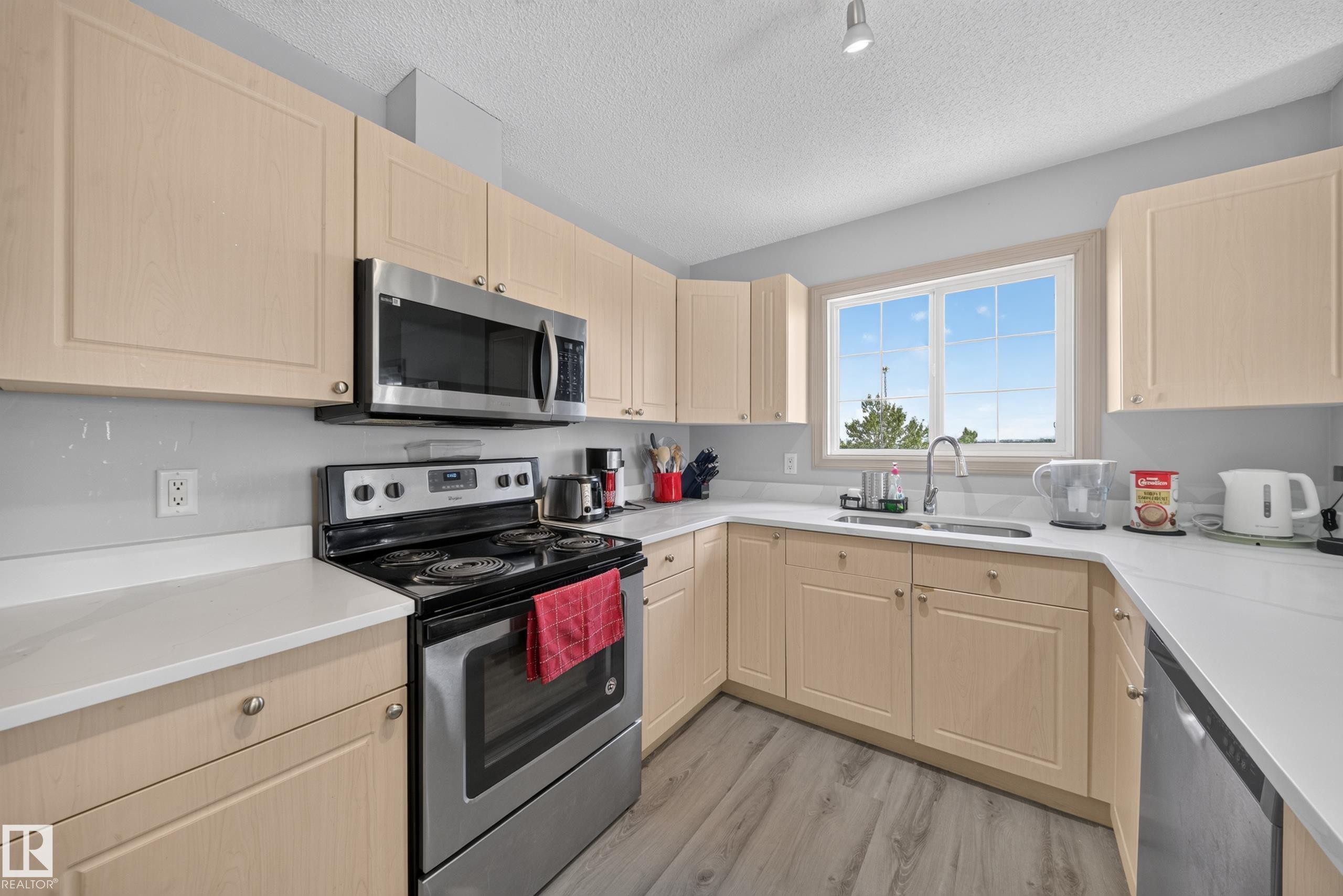 U-shaped kitchen featuring light wood-finish cabinetry, stainless steel appliances, white countertops, and a window above the sink - #419 4407 23 St Nw, Edmonton, AB