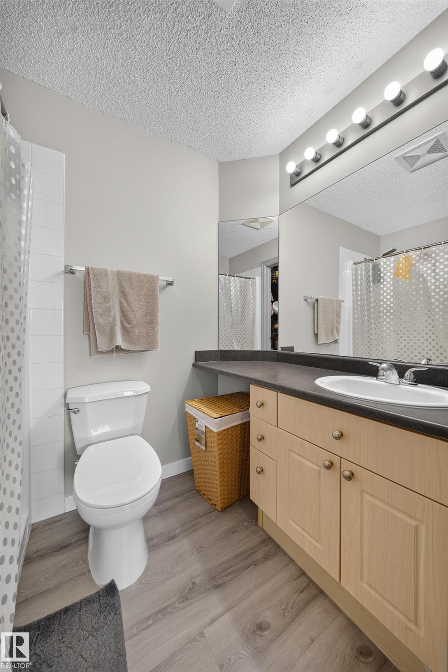 Bathroom featuring wood-finish flooring, a light wood vanity with a dark countertop, a single basin sink, and a large wall-mounted mirror with integrated lighting - #419 4407 23 St Nw, Edmonton, AB