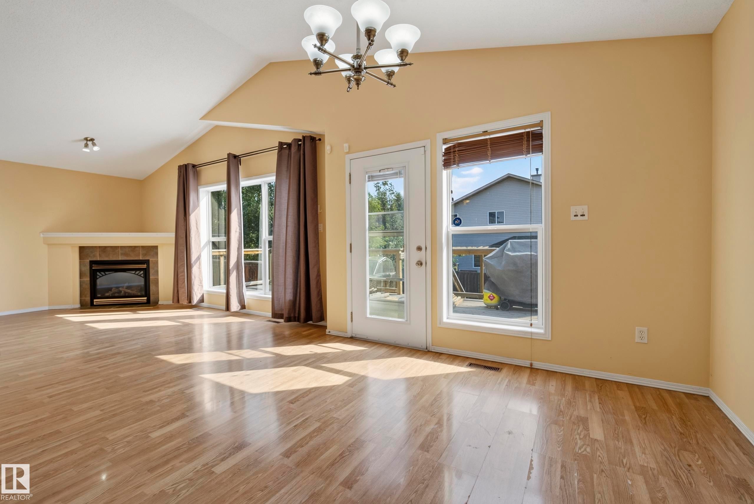 Vaulted ceiling, wood-finish flooring, built-in fireplace with stone-tile surround, multiple windows, and a glass-paneled exterior door - 15213 49A Street Nw, Edmonton, AB