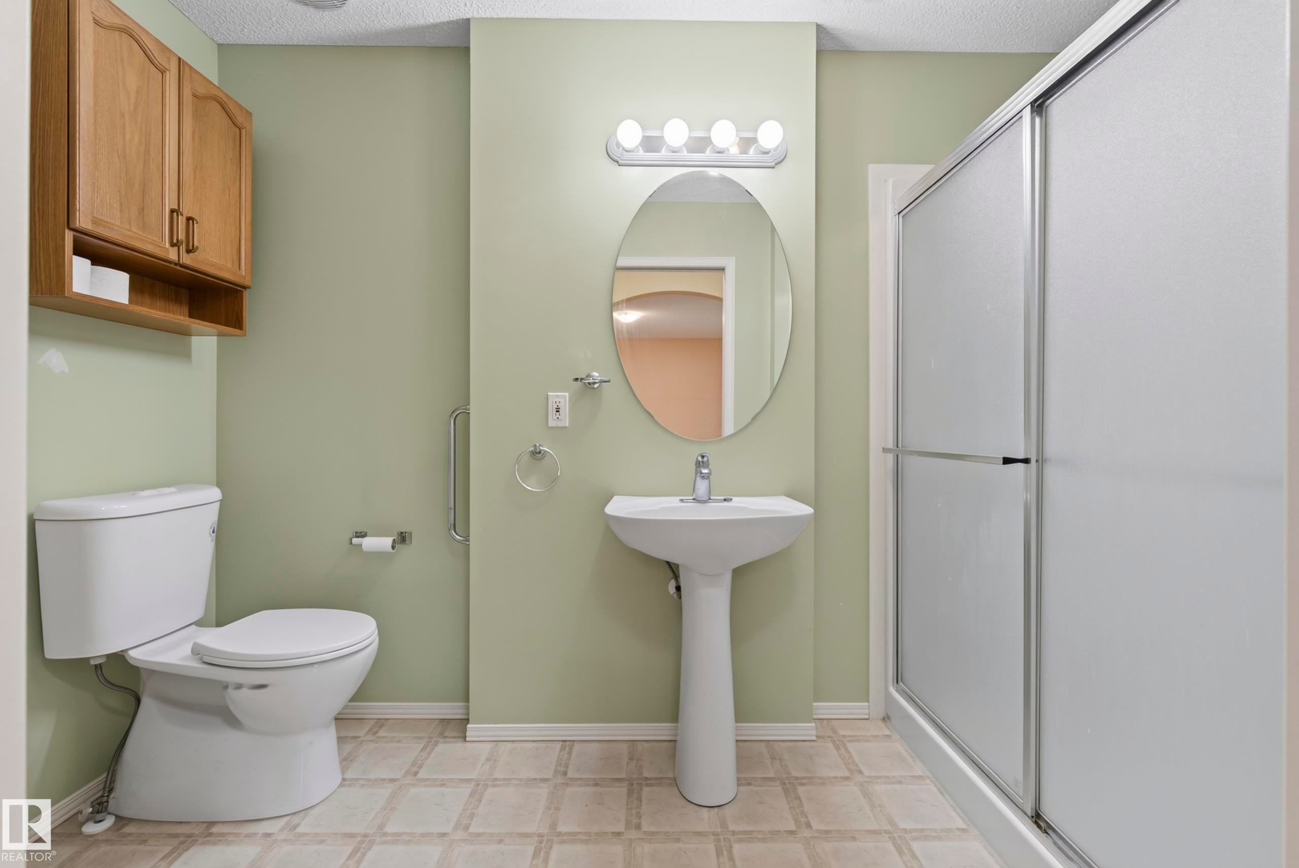Pedestal sink with chrome faucet, oval mirror, shower with sliding frosted glass door, wall-mounted storage cabinet, and tiled flooring - 15213 49A Street Nw, Edmonton, AB