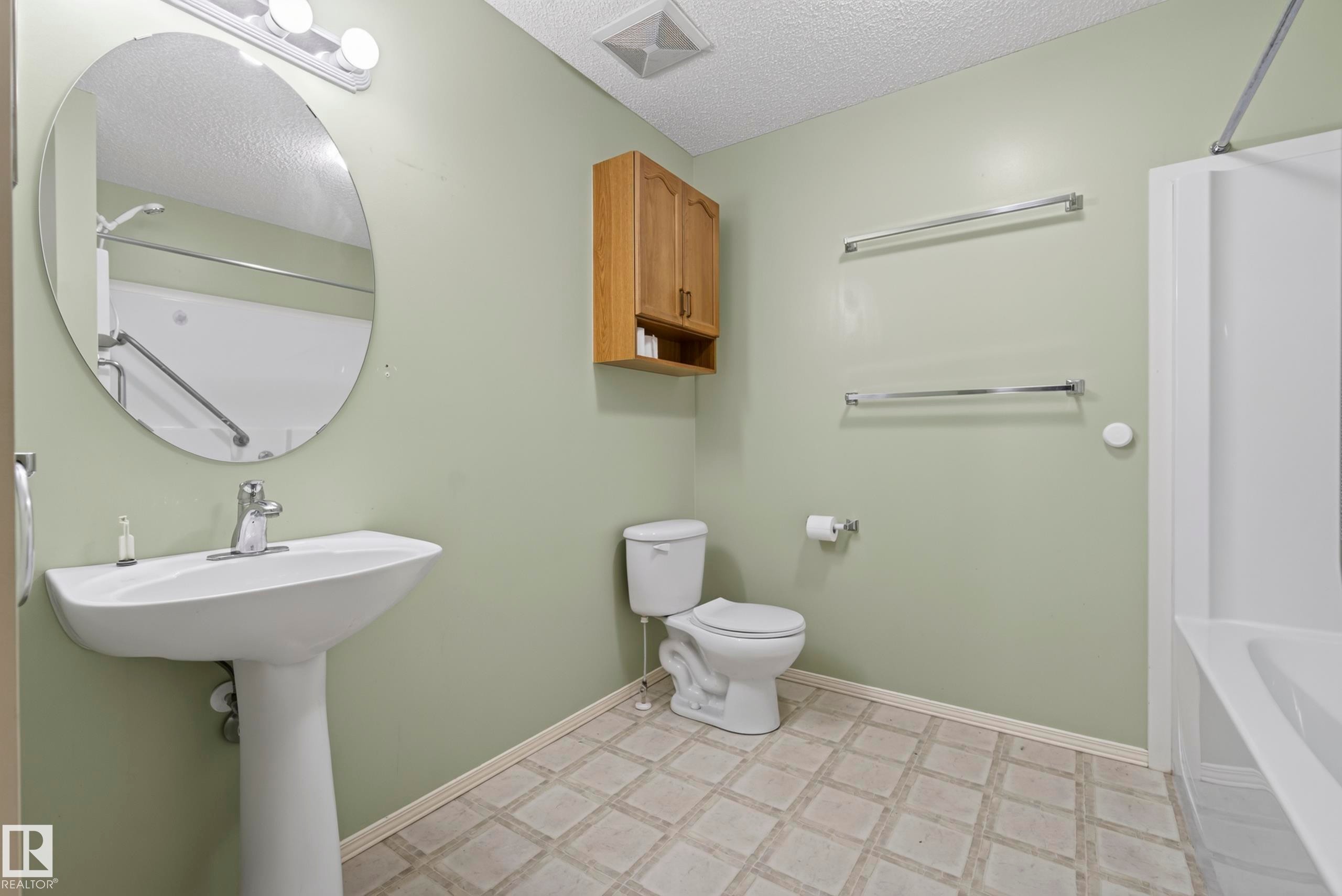 Pedestal sink with chrome faucet, oval mirror, wall-mounted light fixture, wood-finish wall cabinet, and patterned tile-look flooring - 15213 49A Street Nw, Edmonton, AB