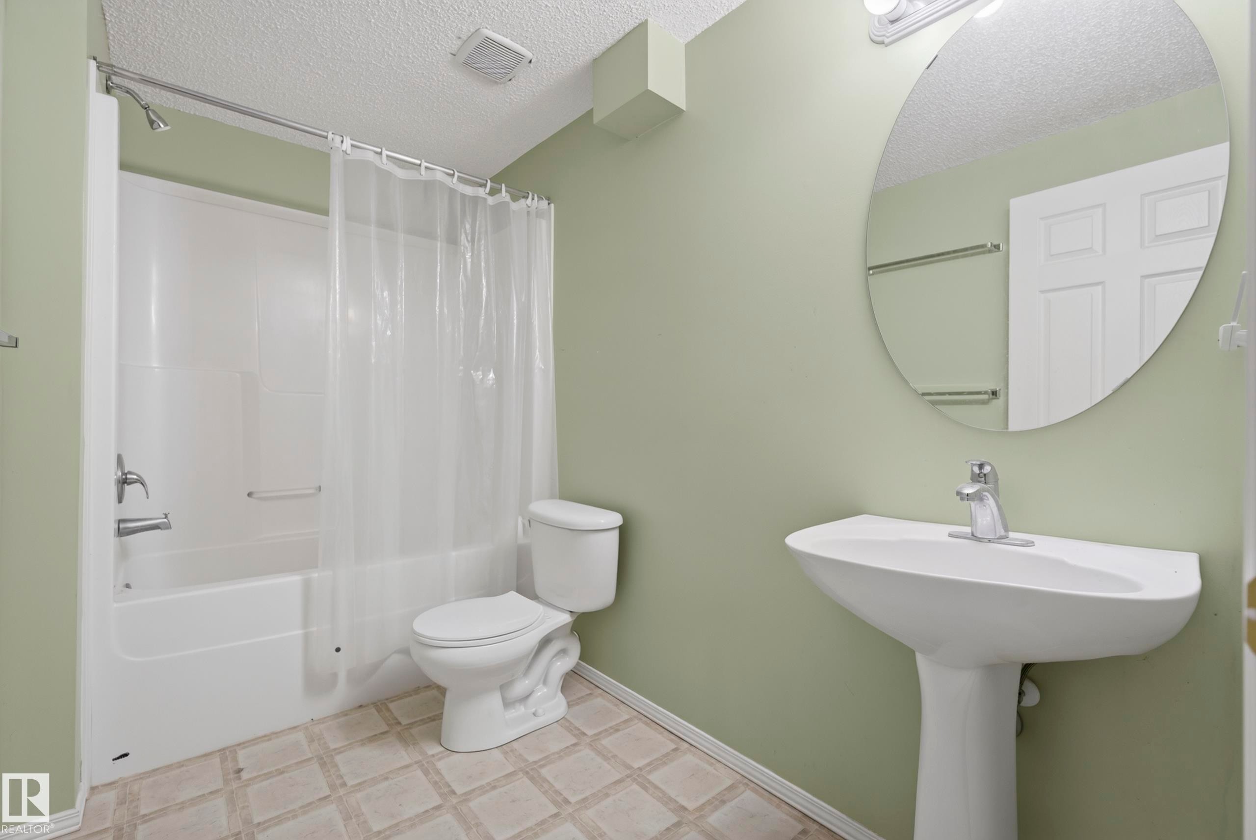 Pedestal sink with chrome faucet, circular wall mirror, and a white bathtub with shower fixtures, set on square-patterned flooring - 15213 49A Street Nw, Edmonton, AB