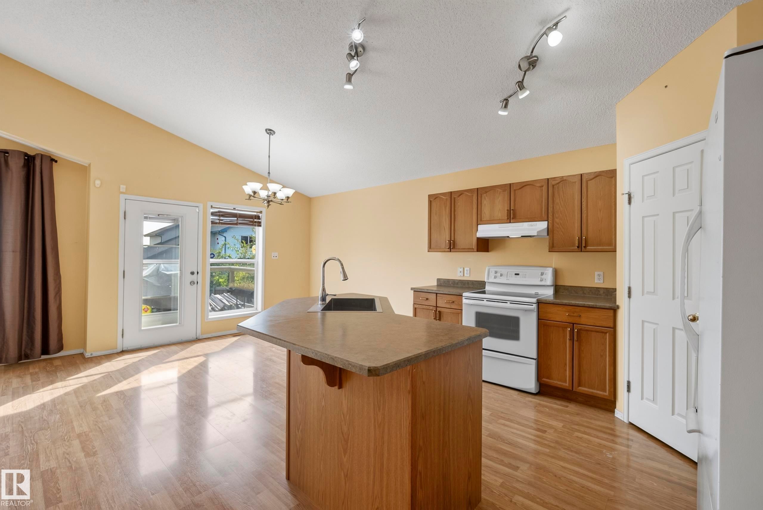 Kitchen featuring wood-finish flooring, a kitchen island with an integrated sink, wood cabinetry, and white appliances - 15213 49A Street Nw, Edmonton, AB
