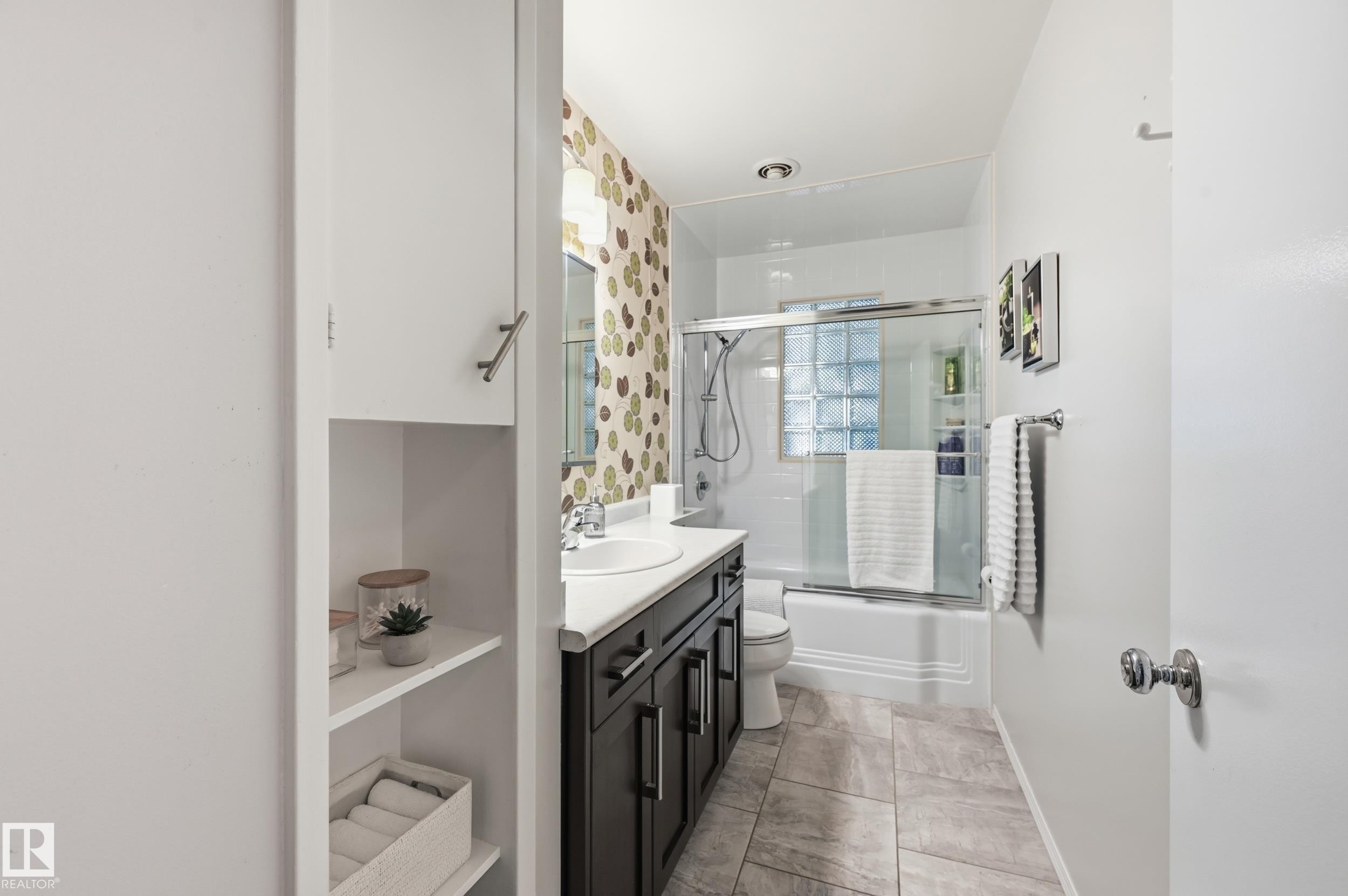 Full bathroom featuring a dark wood-finish vanity with an integrated sink, a glass-enclosed shower-over-tub with glass block accents, and tile flooring - 10552 45 Street Nw, Edmonton, AB
