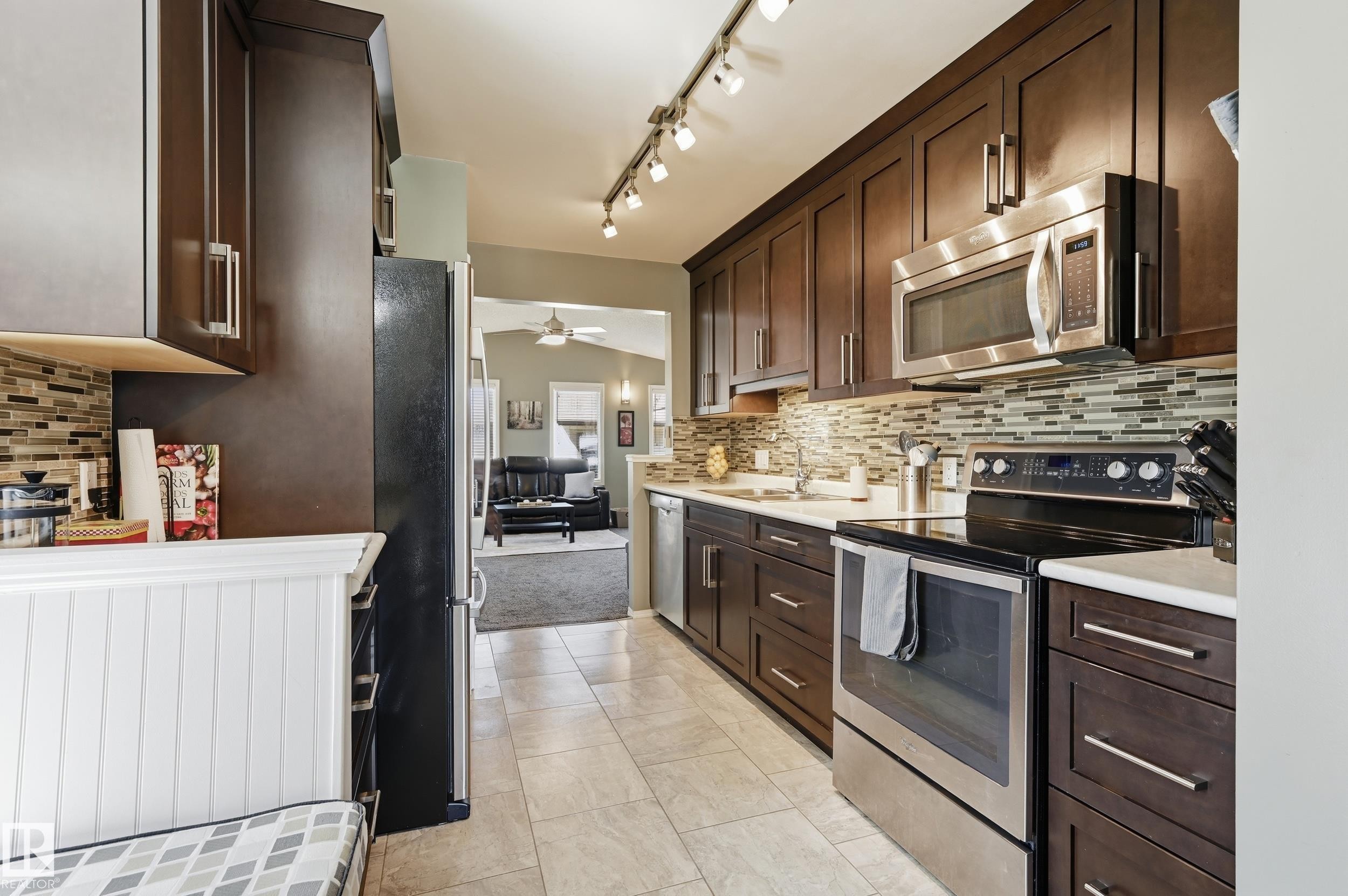 Kitchen featuring wood-finish cabinetry, stainless steel appliances, track lighting, a mosaic tile backsplash, and light-toned flooring - 10552 45 Street Nw, Edmonton, AB