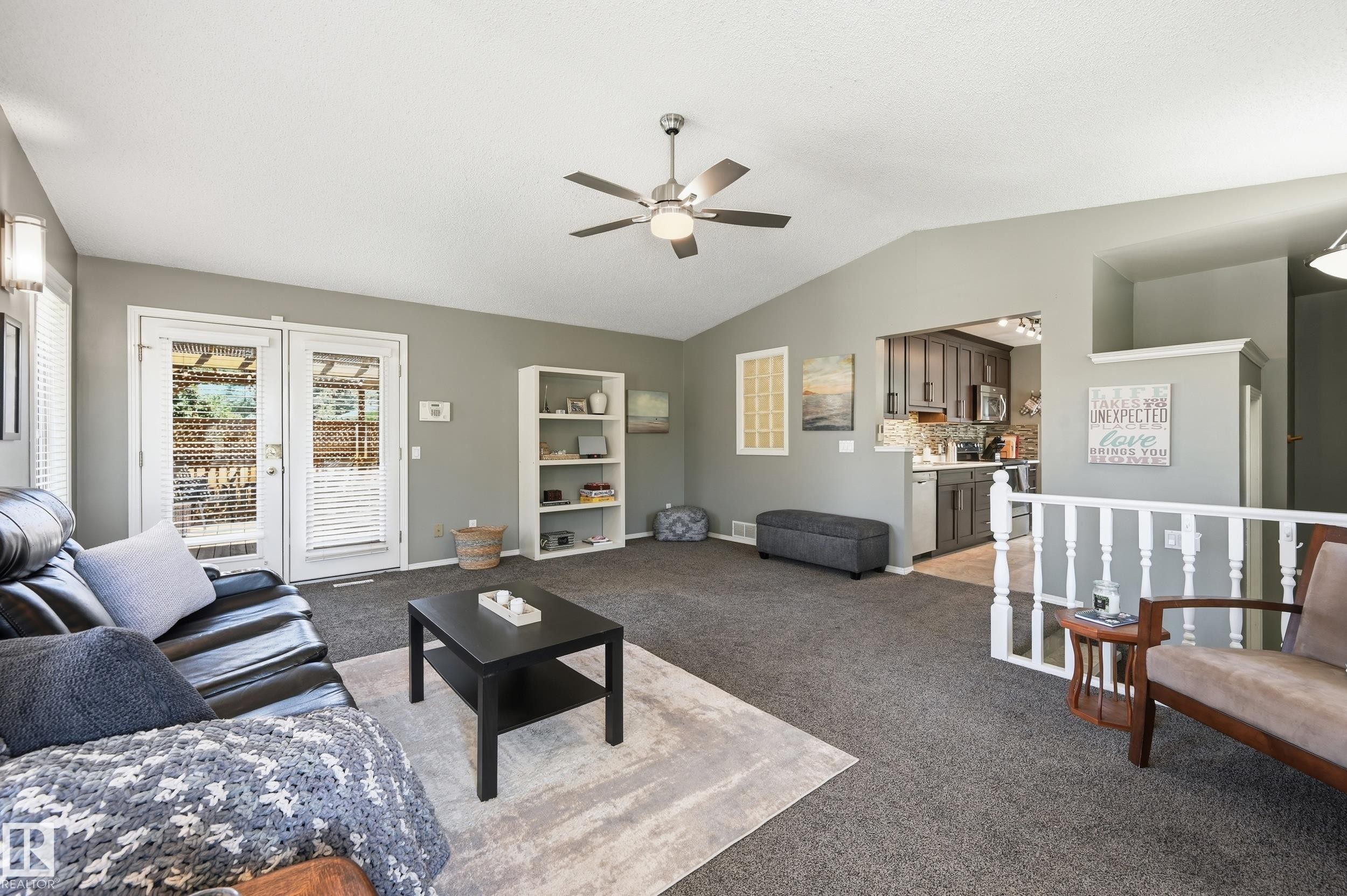 Vaulted ceiling living area with a ceiling fan light fixture, double French doors, and built-in shelving, opening to a kitchen with gray cabinetry and stainless steel appliances - 10552 45 Street Nw, Edmonton, AB