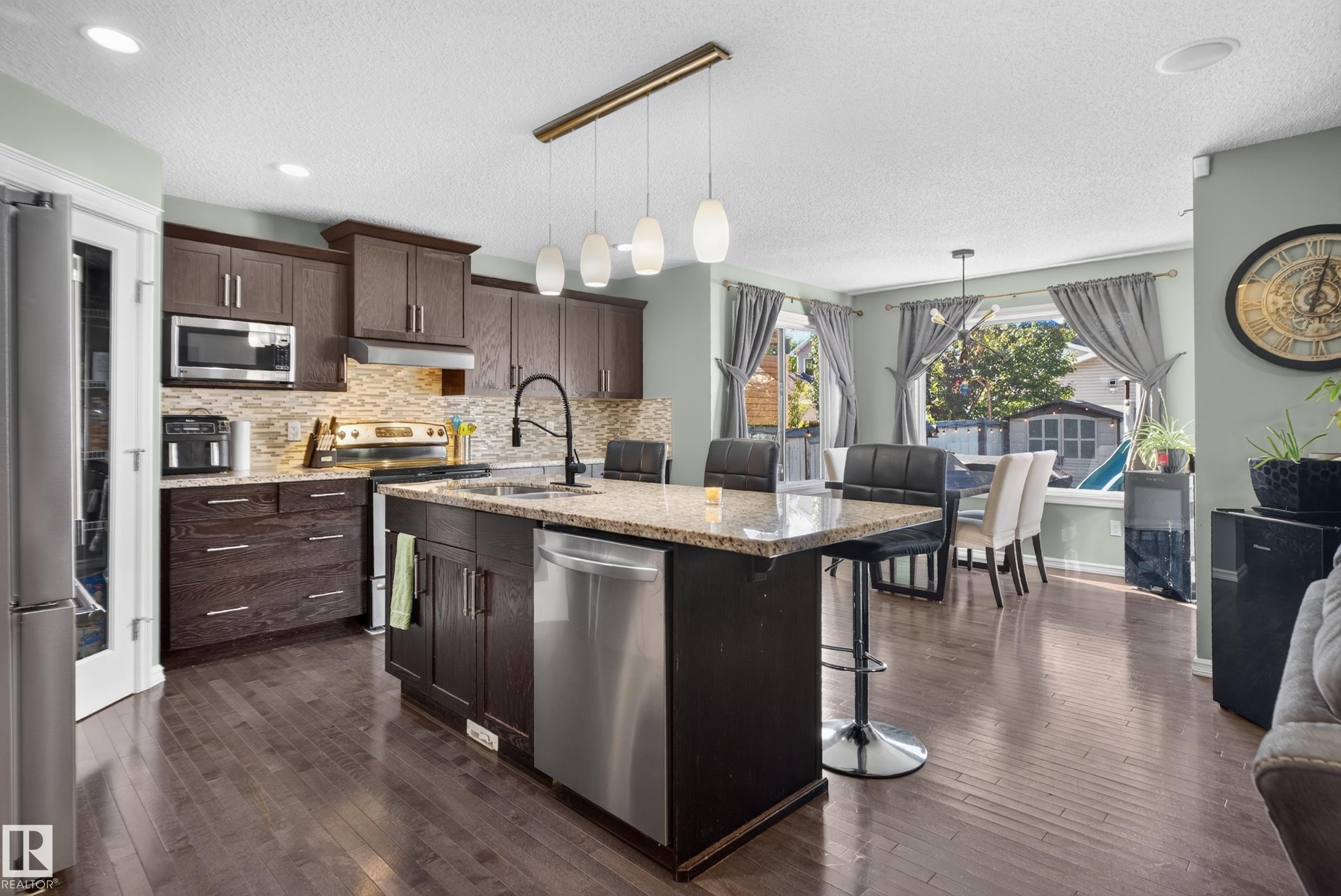 Kitchen featuring dark wood cabinetry, stainless steel appliances, a stone countertop island with seating, a tile backsplash, and recessed lighting - 1332 72 Street Sw, Edmonton, AB