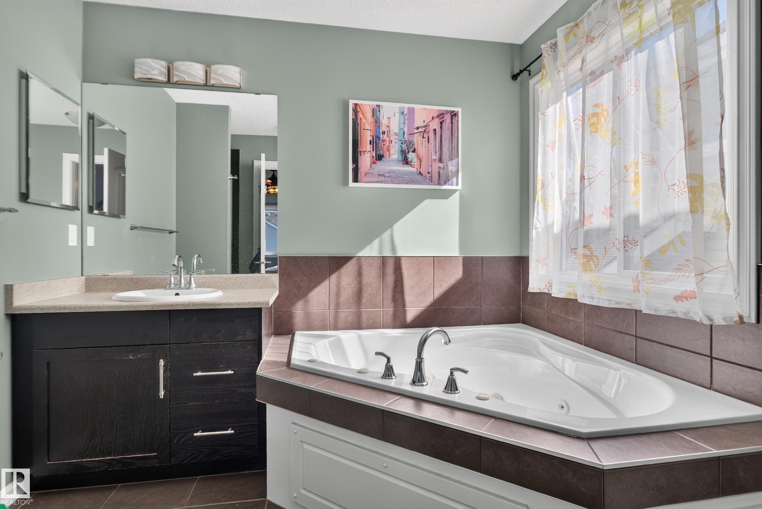 Spa-inspired primary bathroom showcasing a sunken soaking tub with dual faucet fixtures, custom tile surround in warm brown tones, and an expansive vanity with dark cabinetry and quartz countertop - 1332 72 Street Sw, Edmonton, AB