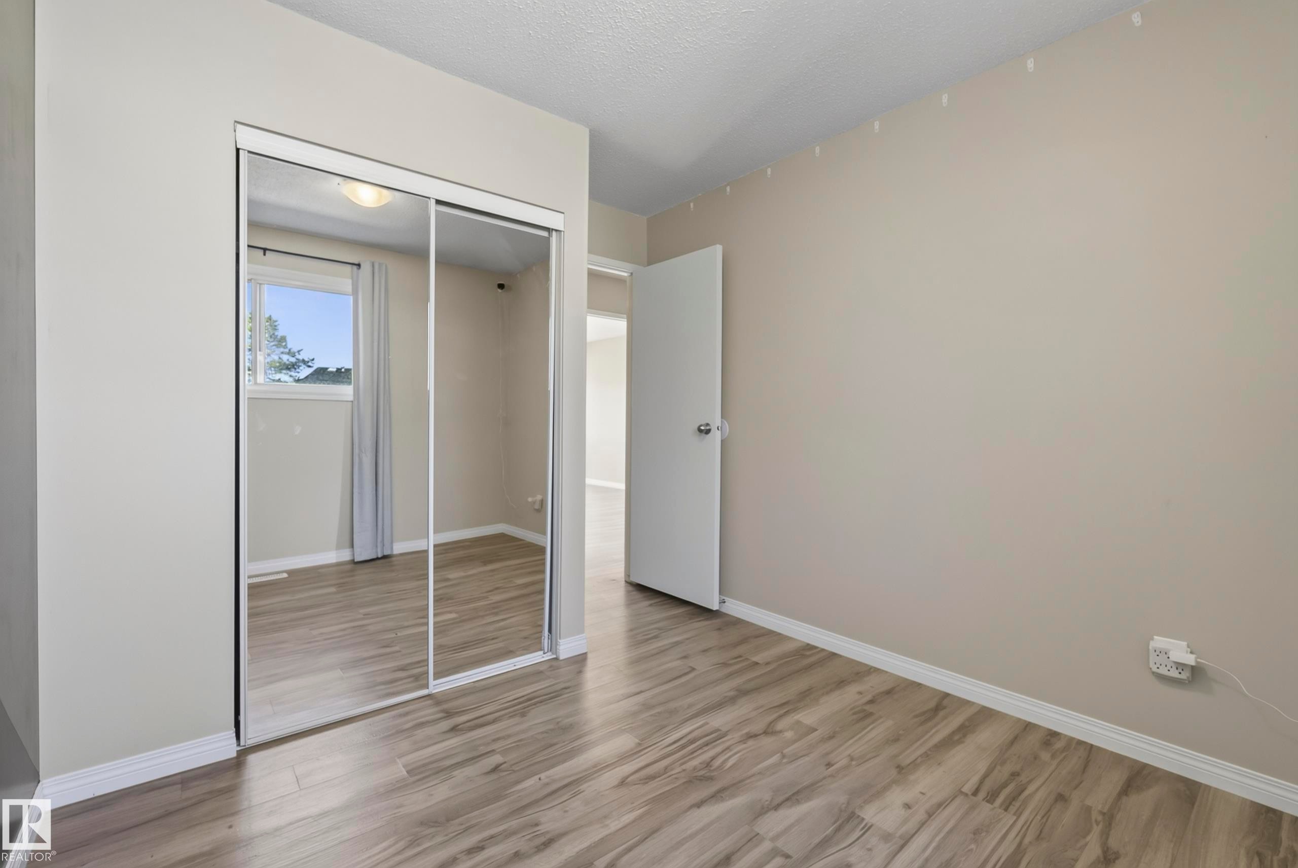 Wood-finish flooring, mirrored sliding closet doors, neutral wall paint, and a bright window with curtain rod - 141 Dickinsfield Court Nw, Edmonton, AB