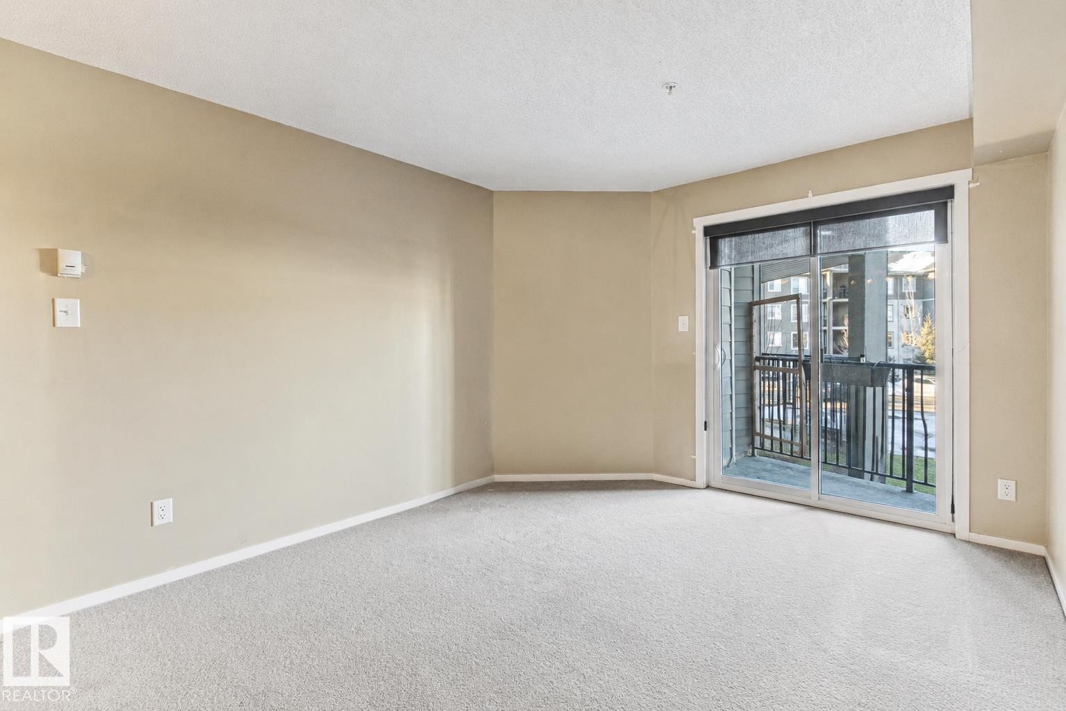 Expansive room with neutral carpeting, light-toned walls, and a sliding glass door leading to an outdoor balcony - #208 18122 77 St Nw, Edmonton, AB