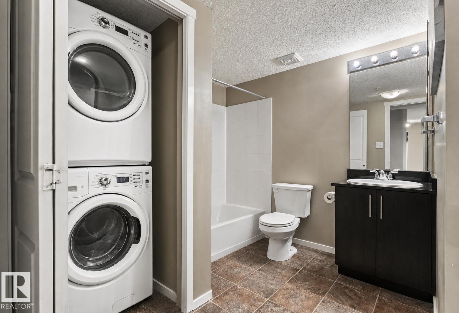 Stacked laundry appliances within a dedicated closet, full bathroom with a dark wood-finish vanity, integrated sink, and overhead light fixture, complete with a bathtub and shower combination - #208 18122 77 St Nw, Edmonton, AB