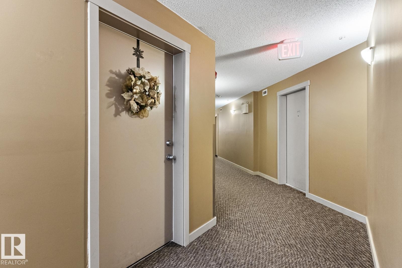 Hallway featuring textured carpet, wall-mounted lighting, and a neutral wall color - #208 18122 77 St Nw, Edmonton, AB