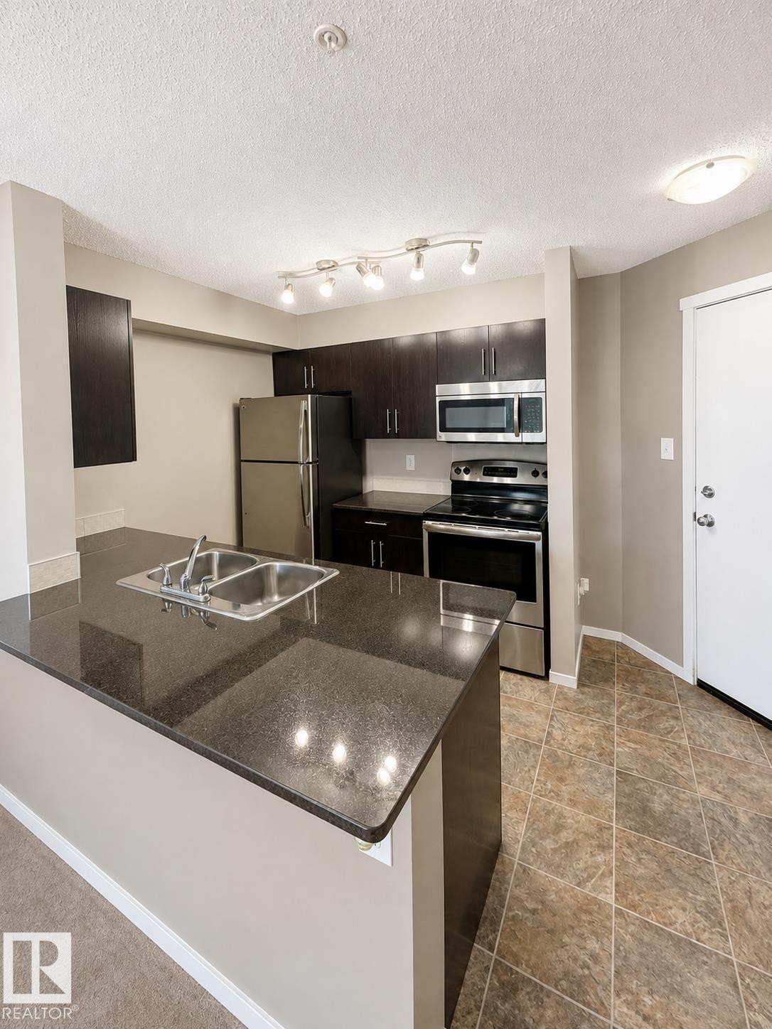 Kitchen featuring dark wood-finish cabinetry, stainless steel appliances, a double basin sink, and a dark stone-finish countertop island - #208 18122 77 St Nw, Edmonton, AB