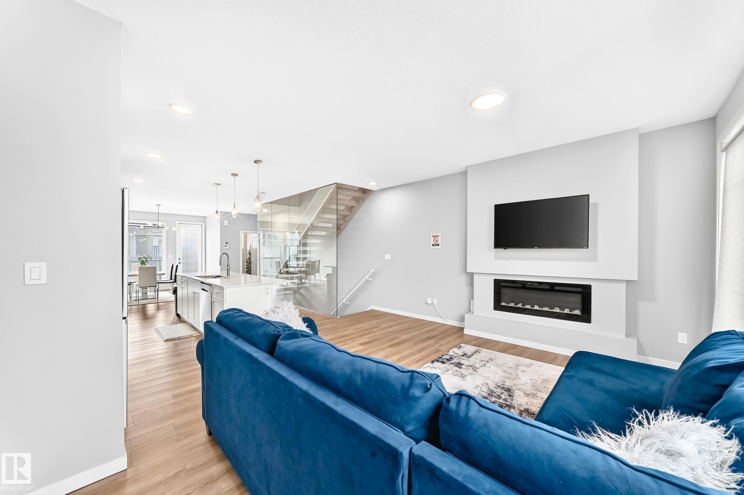 Open-concept living space with wood-finish flooring, a contemporary fireplace feature wall, recessed lighting, a kitchen island with pendant lighting, and a glass-paneled staircase - 479 39 St Sw, Edmonton, AB