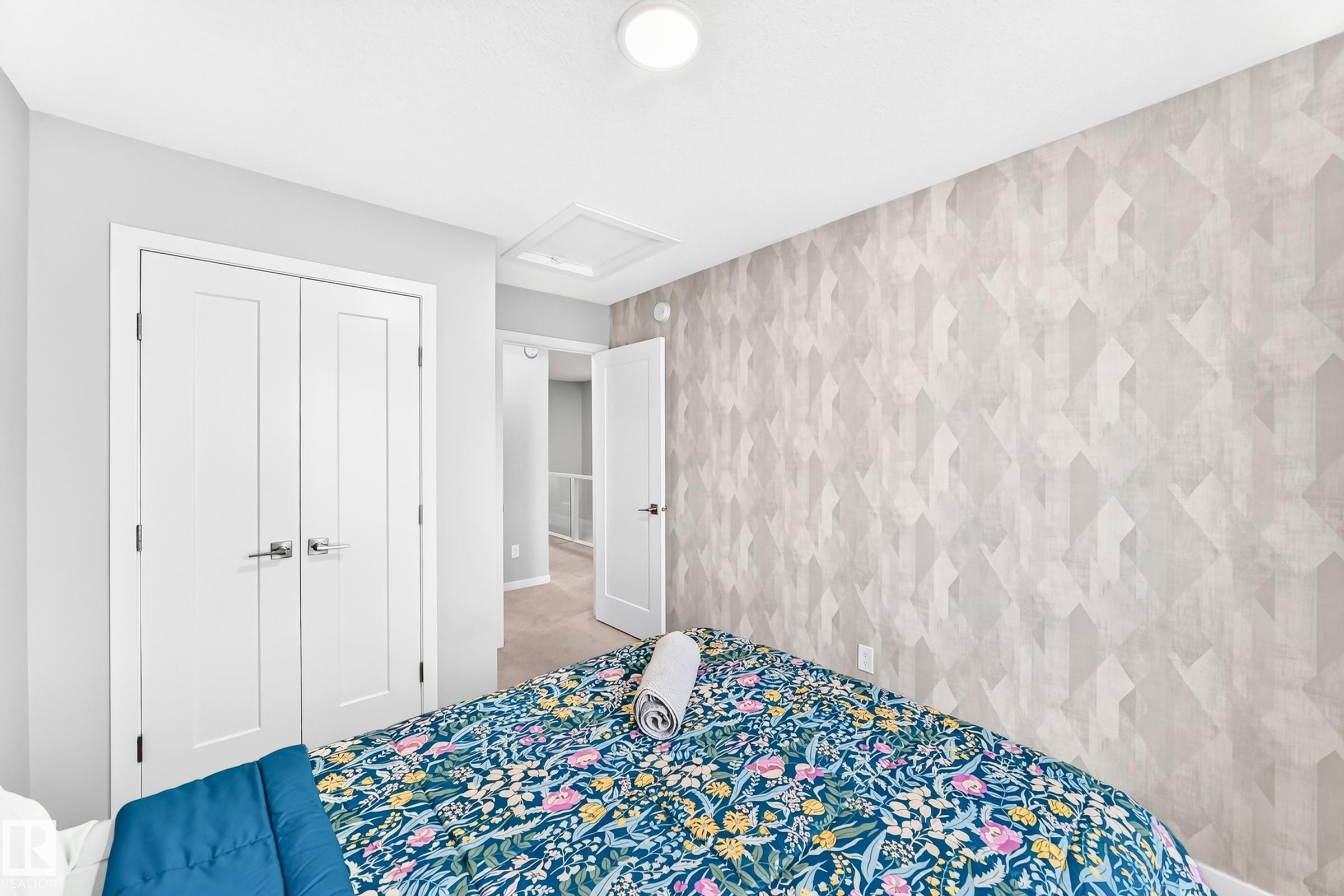 Room featuring a geometric patterned accent wall, white flat-panel closet doors with chrome hardware, an overhead flush-mount light, and light-colored carpeting - 479 39 St Sw, Edmonton, AB