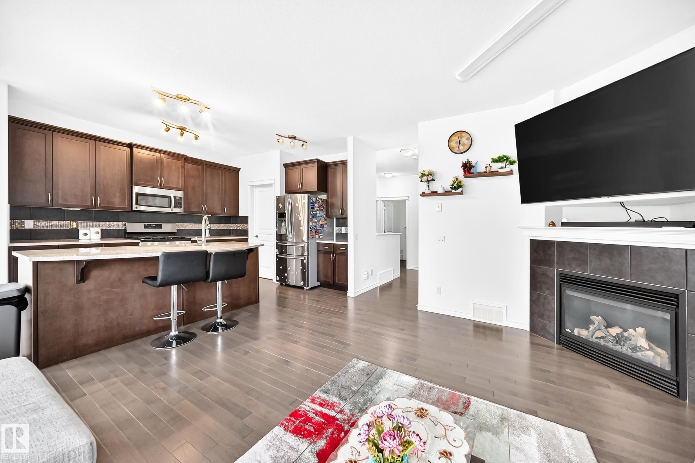 Open-concept living space with wood-finish flooring, a kitchen island with seating, dark wood cabinetry, a stainless steel refrigerator, and a fireplace with a tiled surround - 2015 Chalmers Way Sw, Edmonton, AB