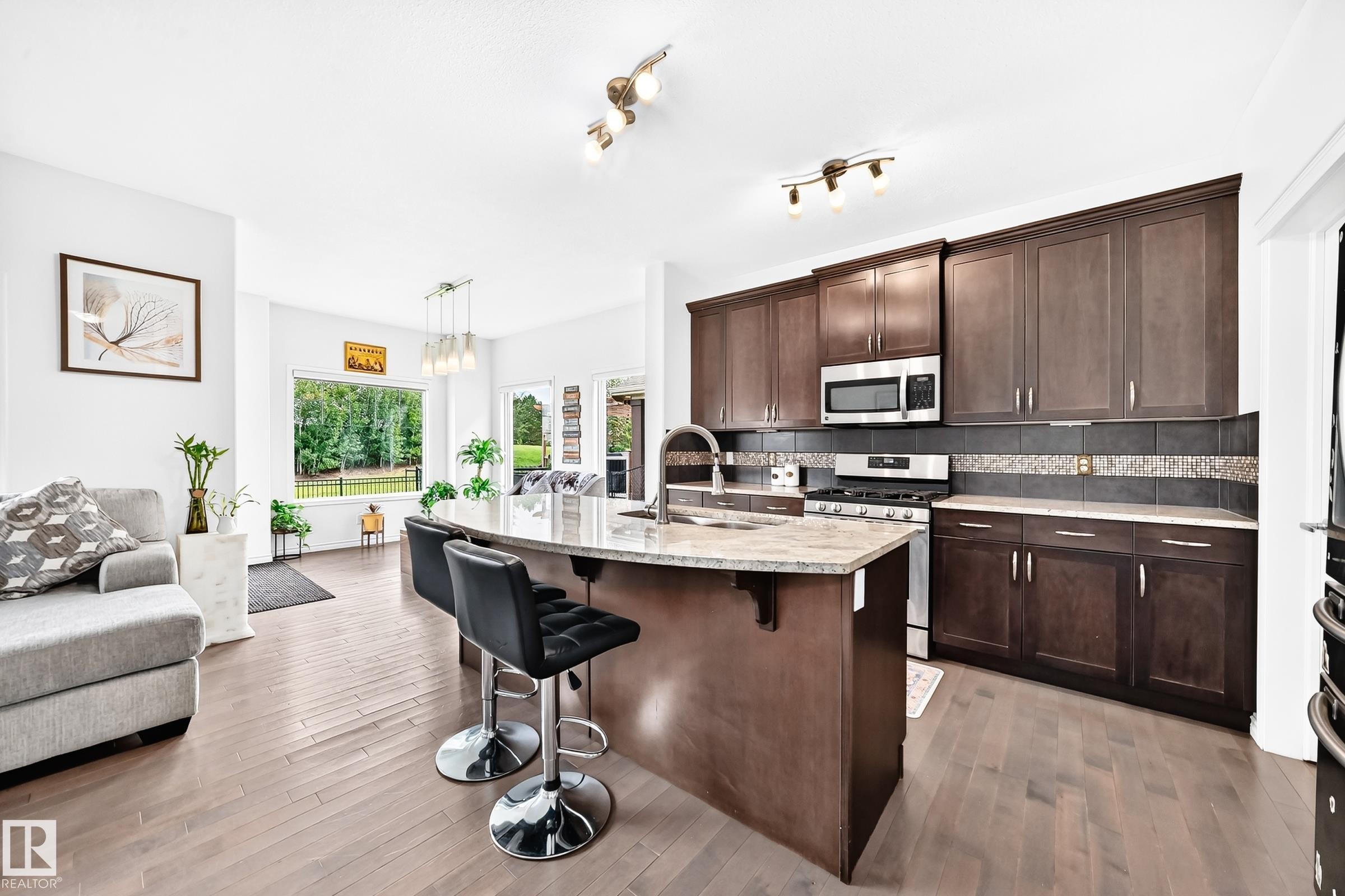 Open-concept living area featuring wood-finish flooring, a kitchen island with stone countertop, stainless steel appliances, dark wood-finish cabinetry, and multiple windows - 2015 Chalmers Way Sw, Edmonton, AB