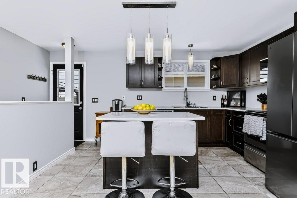Kitchen featuring dark wood-finish cabinetry, light-toned tile flooring, a central island with a solid surface countertop, and pendant lighting fixtures - 18431 95A Avenue Nw, Edmonton, AB