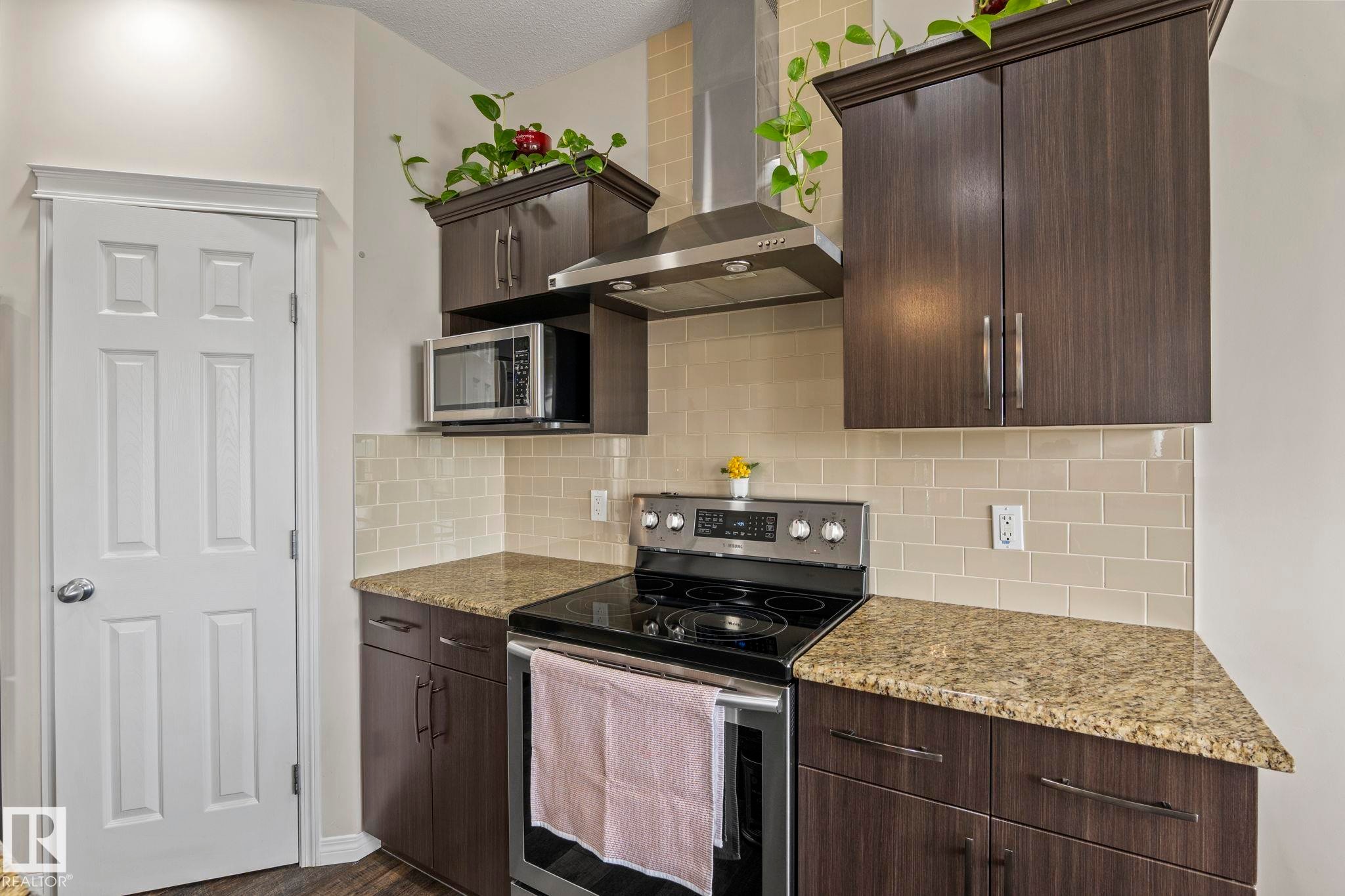 Kitchen featuring dark wood-finish cabinetry, light-toned subway tile backsplash, speckled countertops, stainless steel appliances, and a white paneled door - 2131 51 Sw, Edmonton, AB - Indoor Photo Showing Kitchen