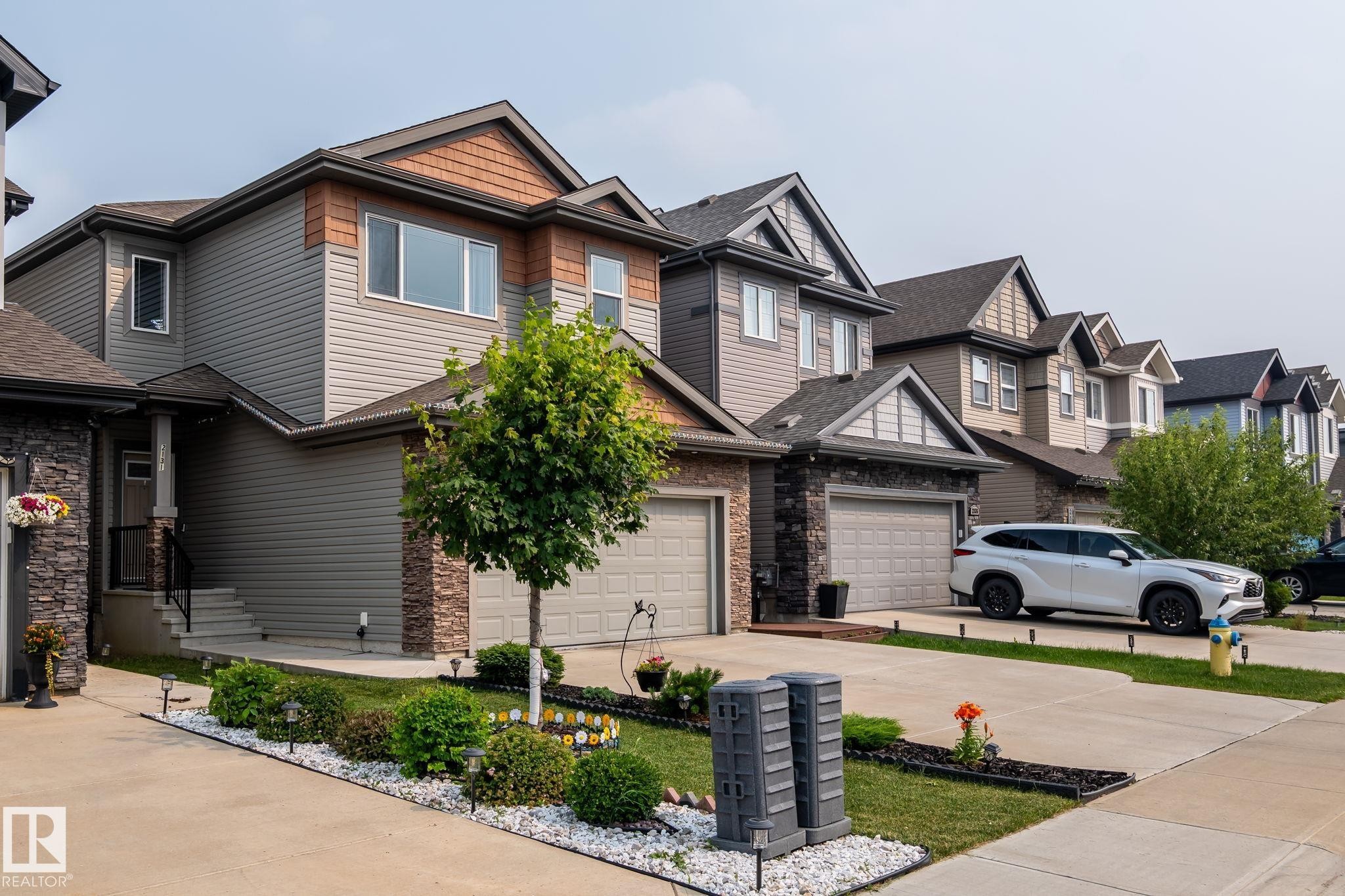 Modern residential exterior featuring gray siding, brown shake accents, stone veneer detailing, a two-car garage, and landscaped front yard with concrete driveway - 2131 51 Sw, Edmonton, AB - Outdoor With Facade
