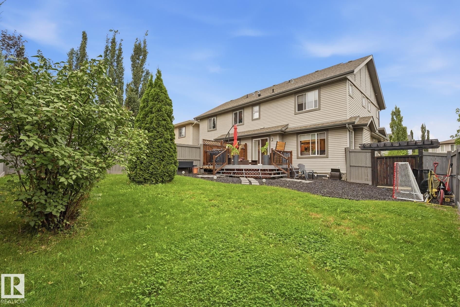 Expansive grassy yard with mature shrubs and trees, leading to a multi-level wood deck and a dedicated gravel area with a wooden pergola - 20007 131 Avenue Nw, Edmonton, AB