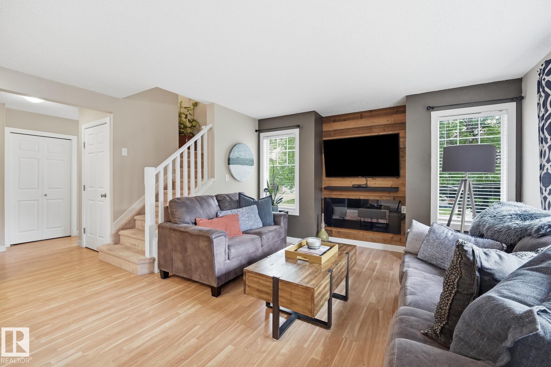 Wood-finish flooring, staircase with white banister, wood-paneled accent wall, built-in electric fireplace, and multiple windows with blinds - 20007 131 Avenue Nw, Edmonton, AB