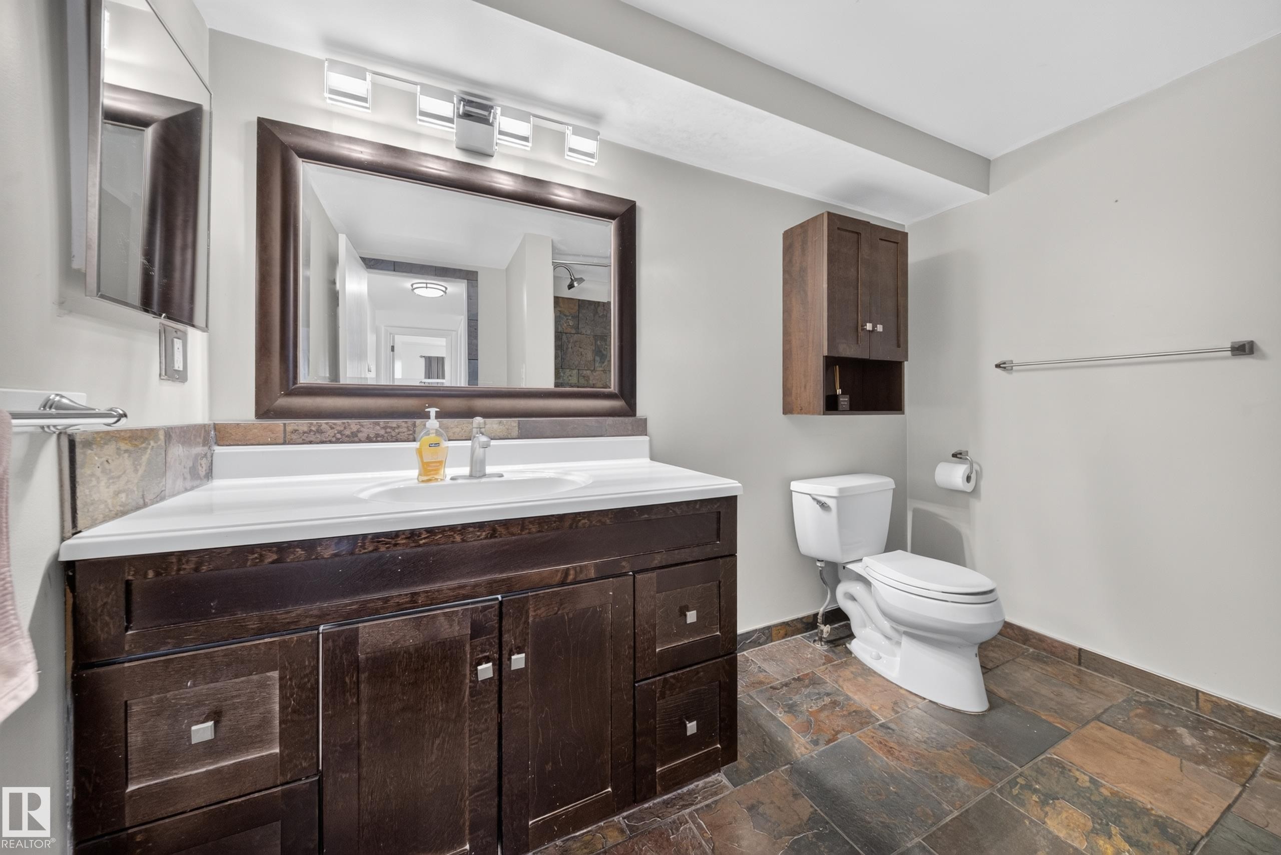 Dark wood-finish vanity with integrated sink, stone tile flooring, framed mirror, and wall-mounted cabinetry - 155 Marlborough Place Nw, Edmonton, AB