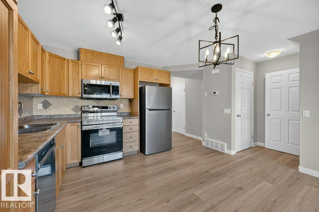 Kitchen featuring wood-finish cabinetry, stainless steel appliances, patterned tile backsplash, wood-look flooring, and a decorative chandelier - #26 2816 34 Av Nw, Edmonton, AB