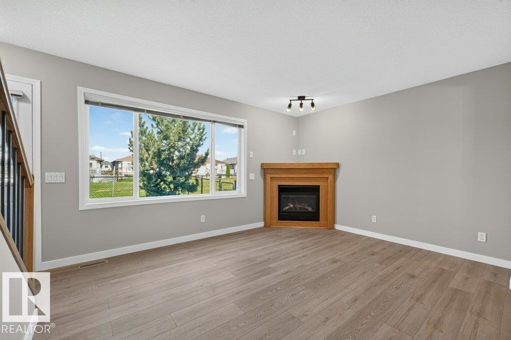Spacious room featuring wood-finish flooring, a contemporary fireplace with a wood mantle, and a large multi-pane window - #26 2816 34 Av Nw, Edmonton, AB